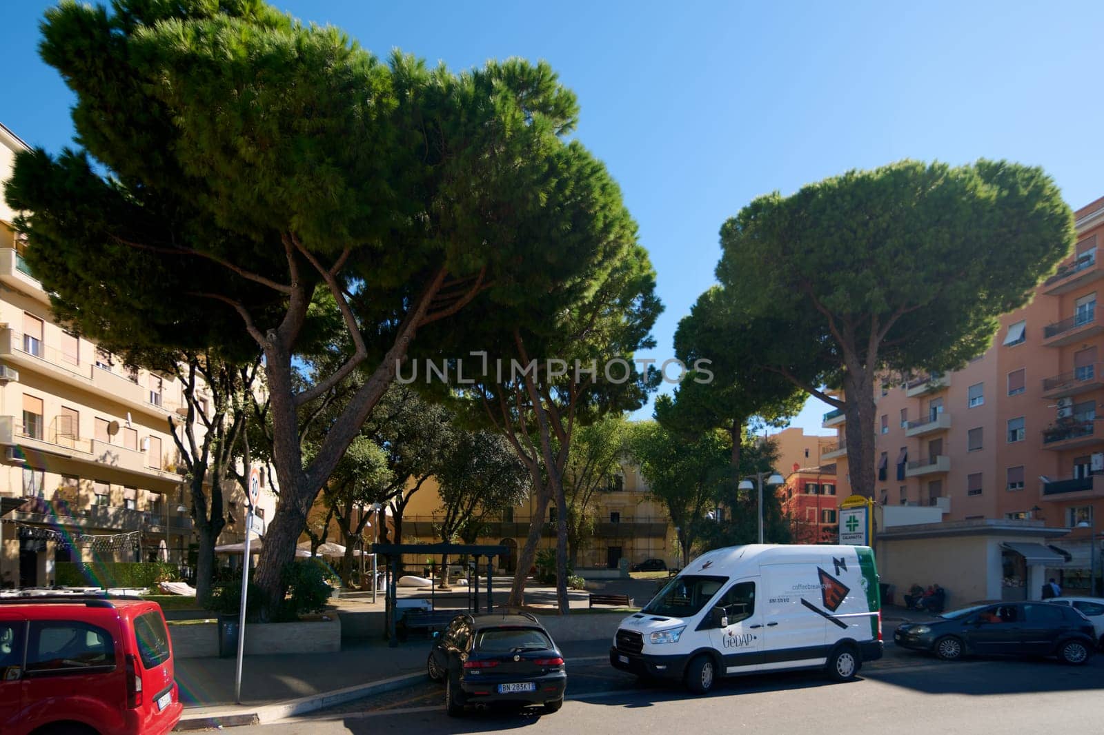 Civitavecchia, Italy - December 19, 2024: Residential square with parked cars, a van. Nearby buildings include apartments and a pharmacy. Concept: Neighborhood square and urban life.