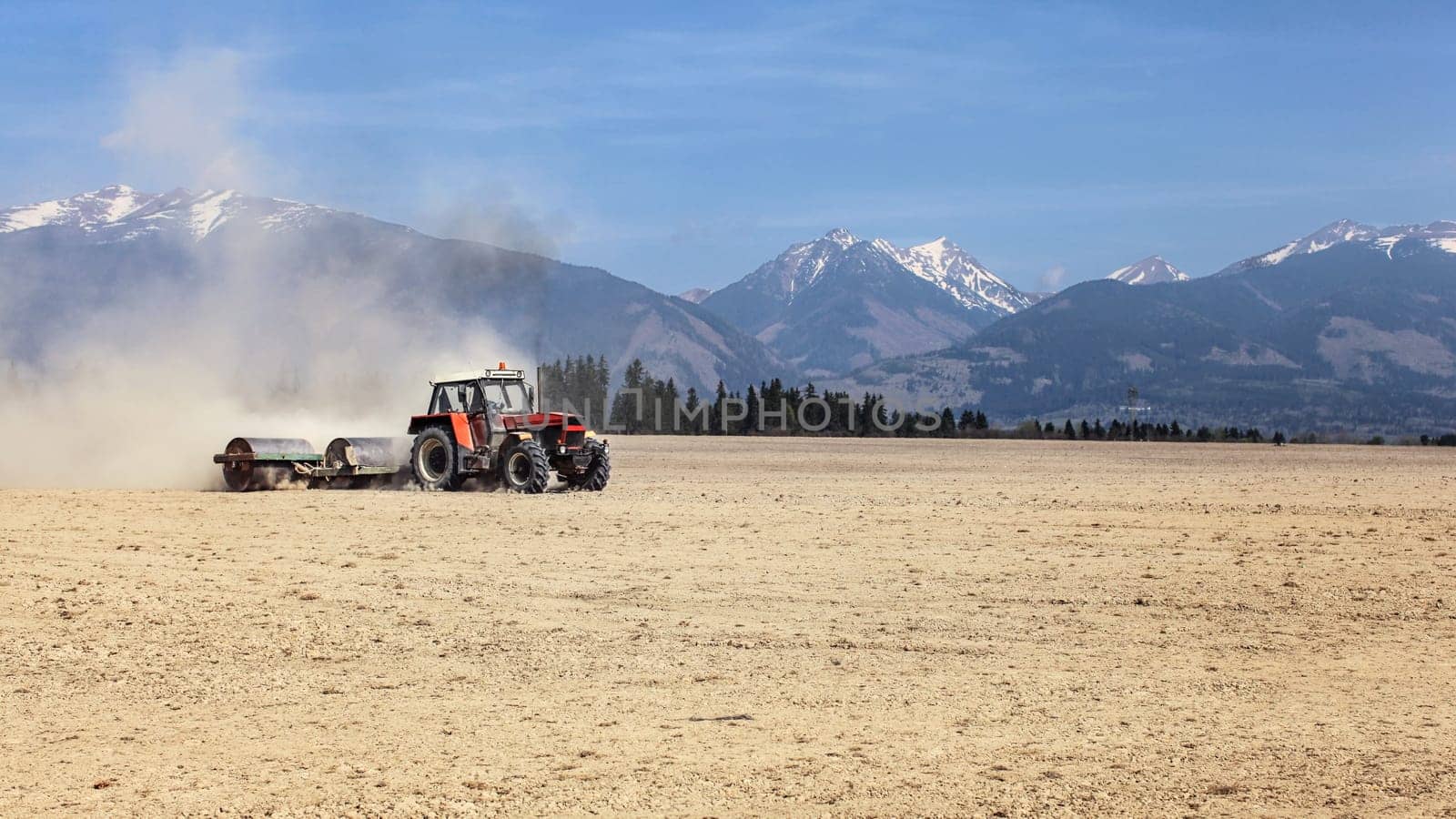 Tractor pulling metal rollers over dry field with mountains in background. Spring soil preparation.