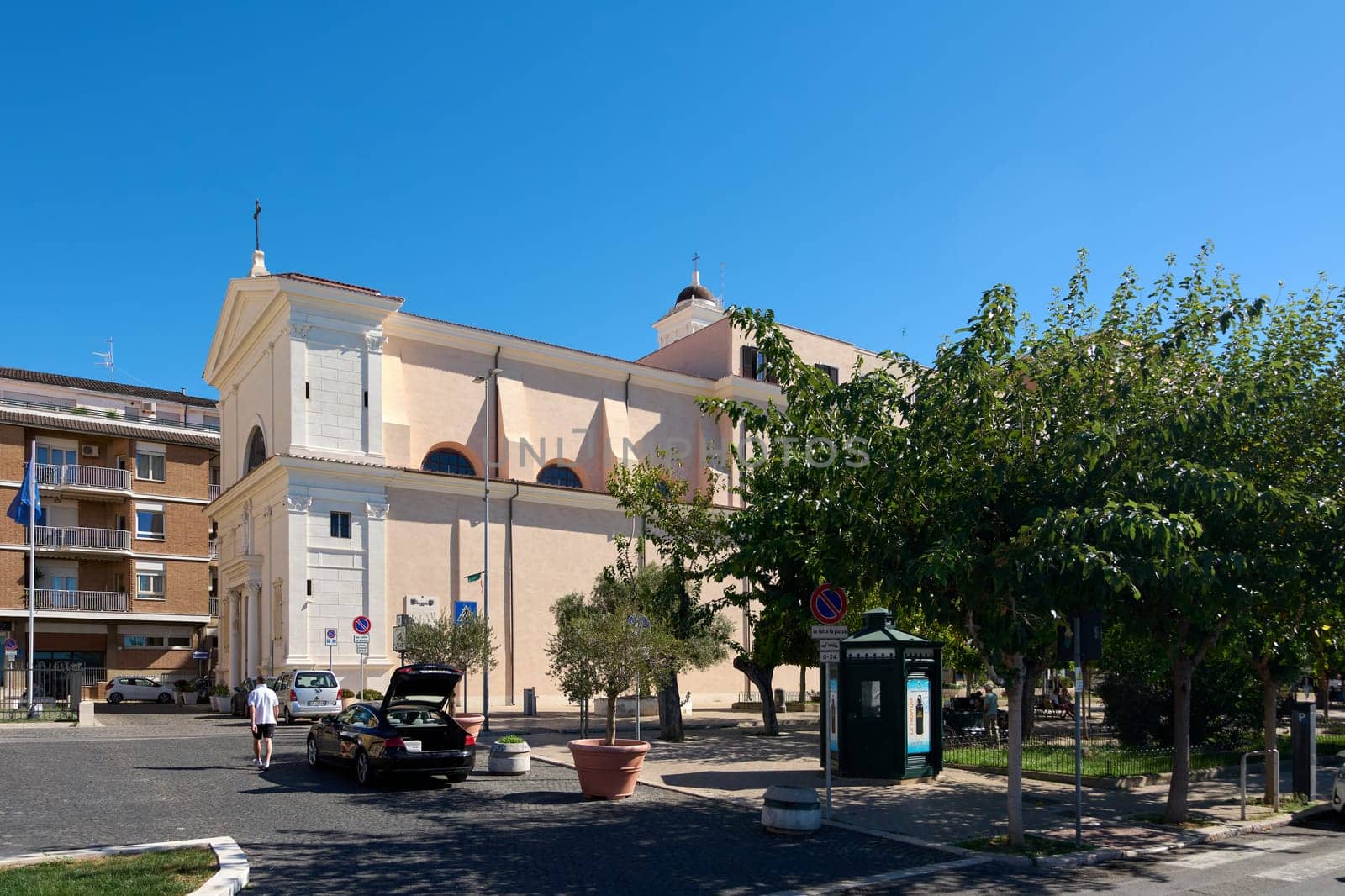 Civitavecchia. Italia  - December 27, 2024: An urban square in Civitavecchia, Italy, with a historical church, trees, and parked cars under a bright sunny sky. Concept: Cityscape and cultural heritage