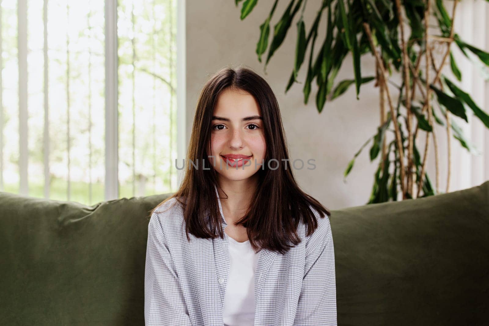 Beautiful smiling caucasian girl sitting at green sofa and watching at camera. Cute woman front view. Female smile, looking in photo camera. Online interview or meeting. Big lips. High quality photo
