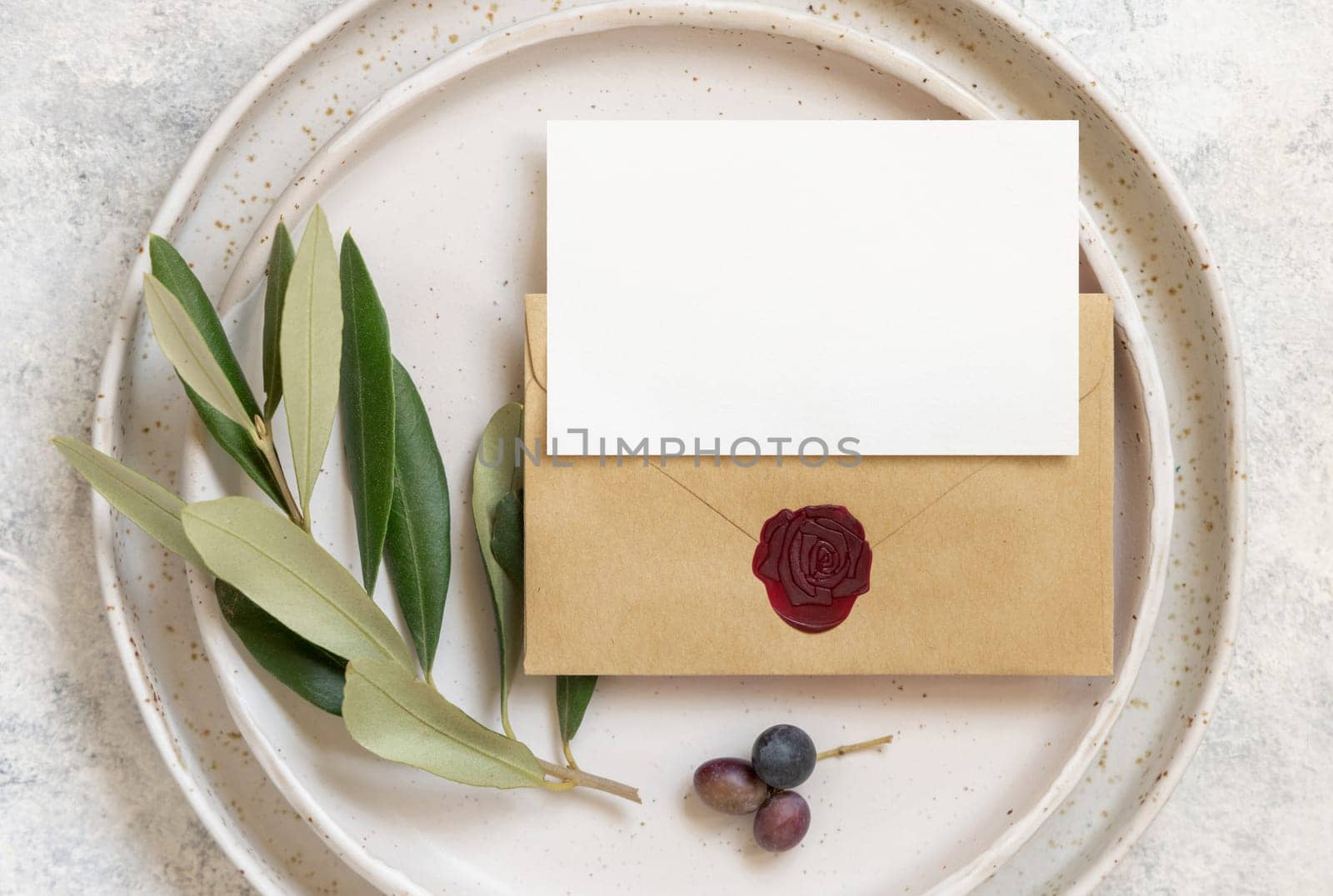Wedding invitation card and envelope over porcelain plates on white marble table with olive branches top view. Elegant modern template with horizontal blank paper card. Mediterranean flat lay mockup
