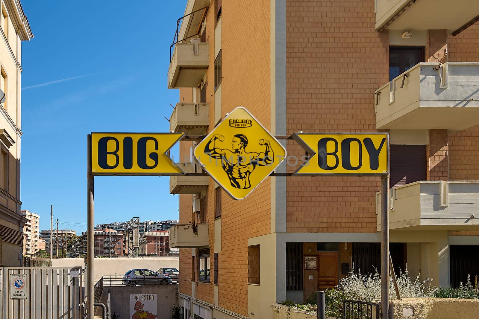 Civitavecchia. Italia - December 27, 2024: Big Boy Gym sign with illustration of muscular man. Mounted on metal structure among buildings, blue sky and parked cars, urban fitness center concept