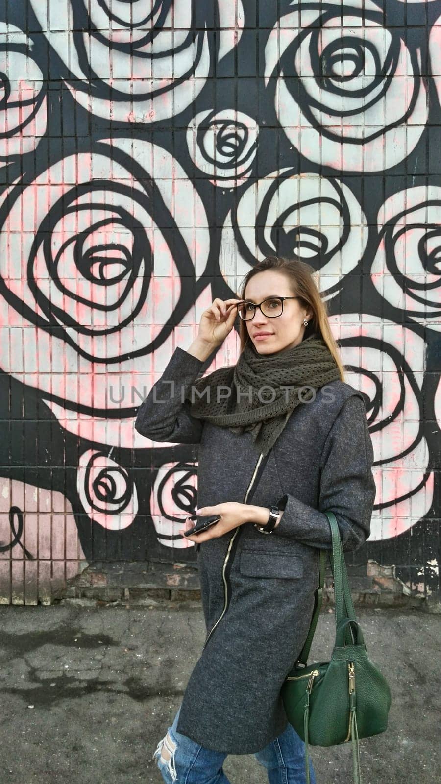 A woman in a grey coat and green scarf with a green bag puts on glasses