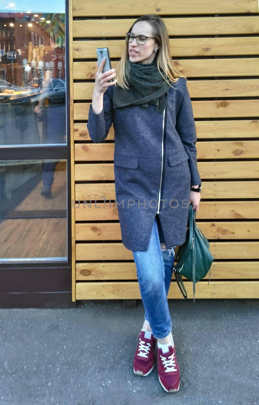 A young woman in a coat and jeans indignantly looks at the smartphone screen in the street against the background of a wooden wall