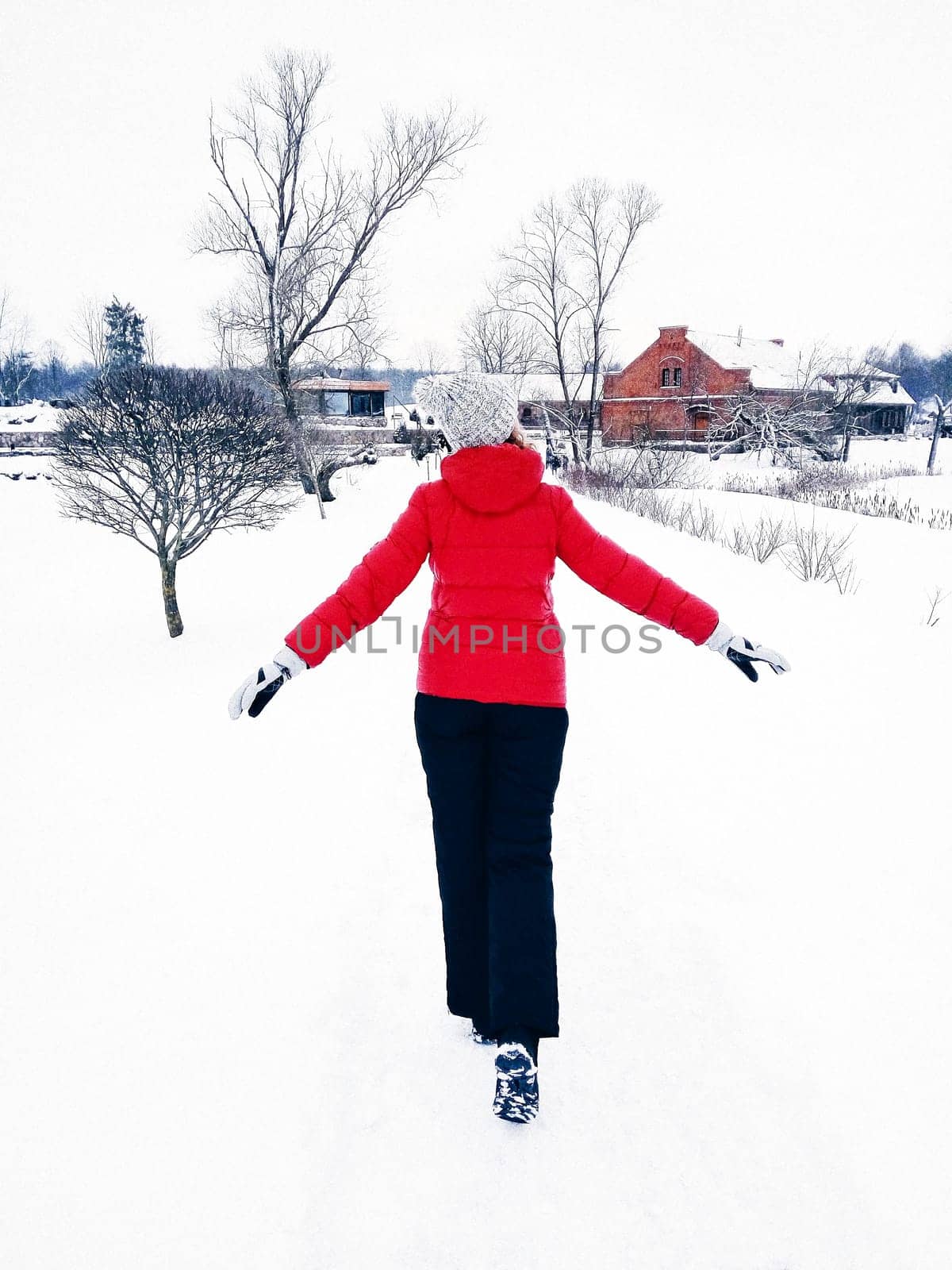 View from the back of a woman in a red jacket, a white hat and black sweatpants walking in the winter through the snow outside the city, winter domestic travel