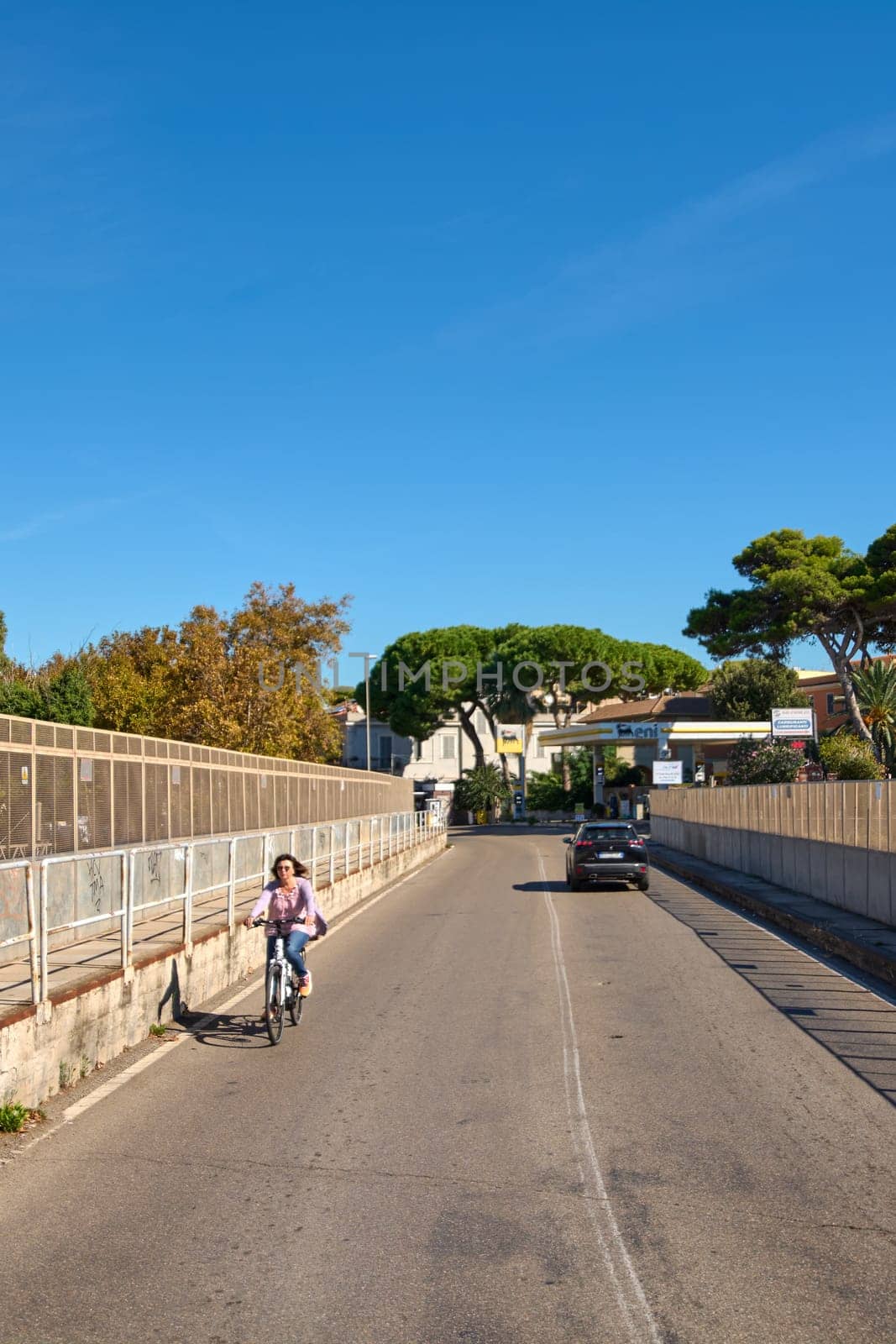 Civitavecchia, Italy - December 27, 2024: Cyclist rides on urban street in Civitavecchia on a sunny day. The road with fences and trees, with a car in the background. Concept: urban transport.