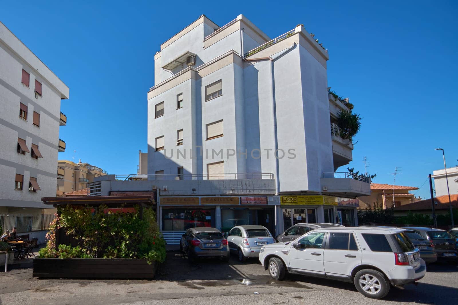 Civitavecchia. Italia - December 27, 2024: Multi-story building with gray facade, windows, and storefronts on ground floor. Parked cars in front and blue sky. Concept: Urban architecture and commerce.