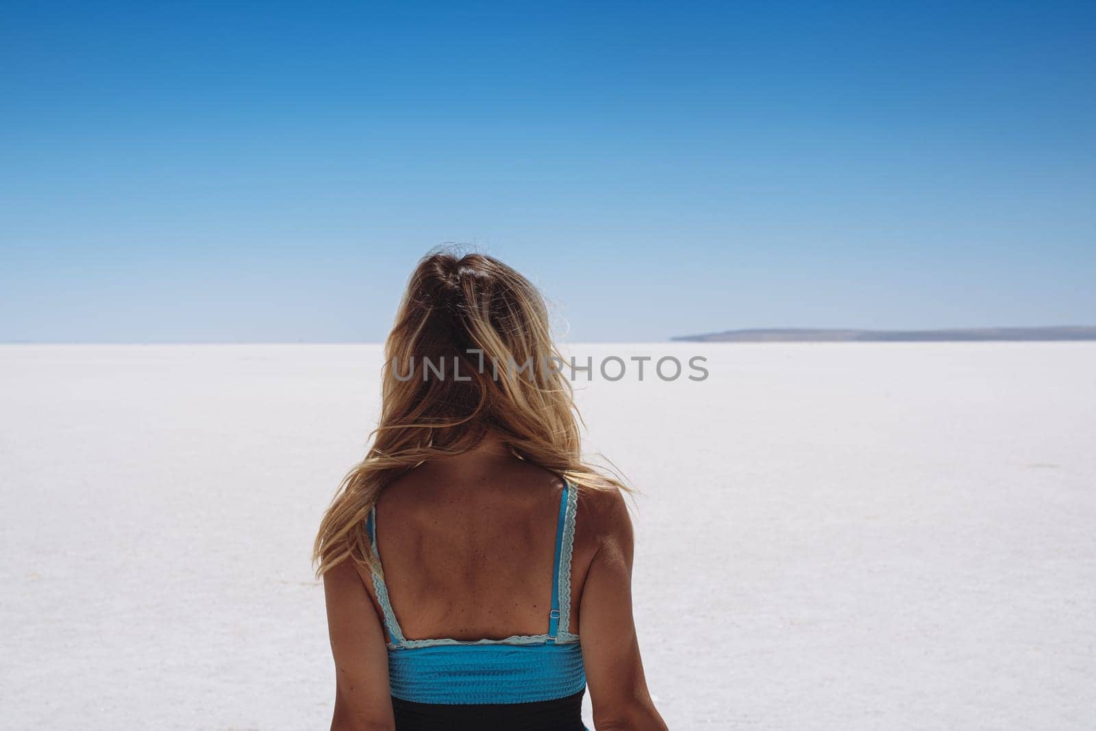 rear view of blonde beautiful Caucasian young woman dressed in blue looking at white salt lake stretching to the horizon with clear sky above