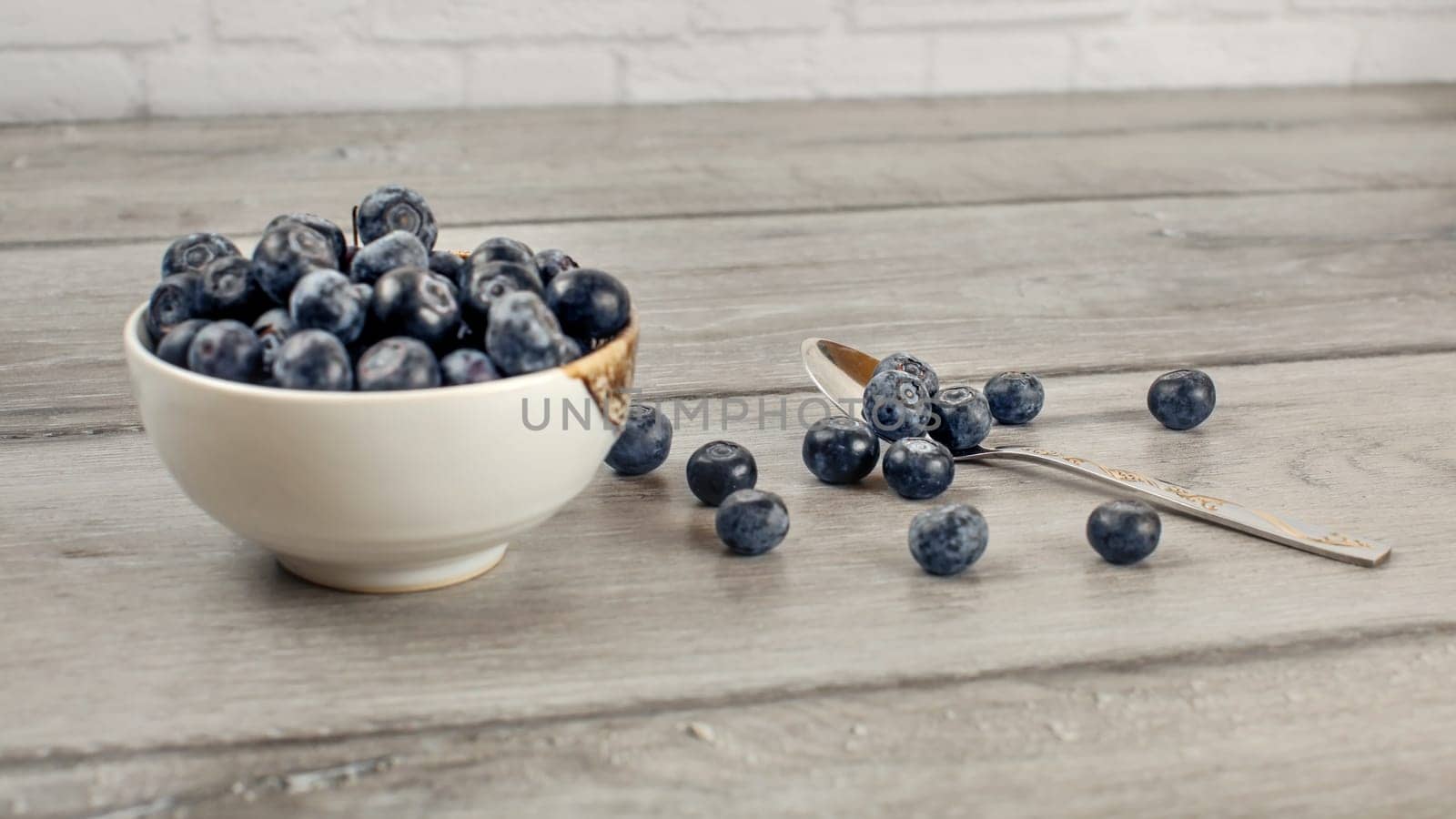 Small bowl of blueberries, some of them spilled next to spoon on gray wood desk.