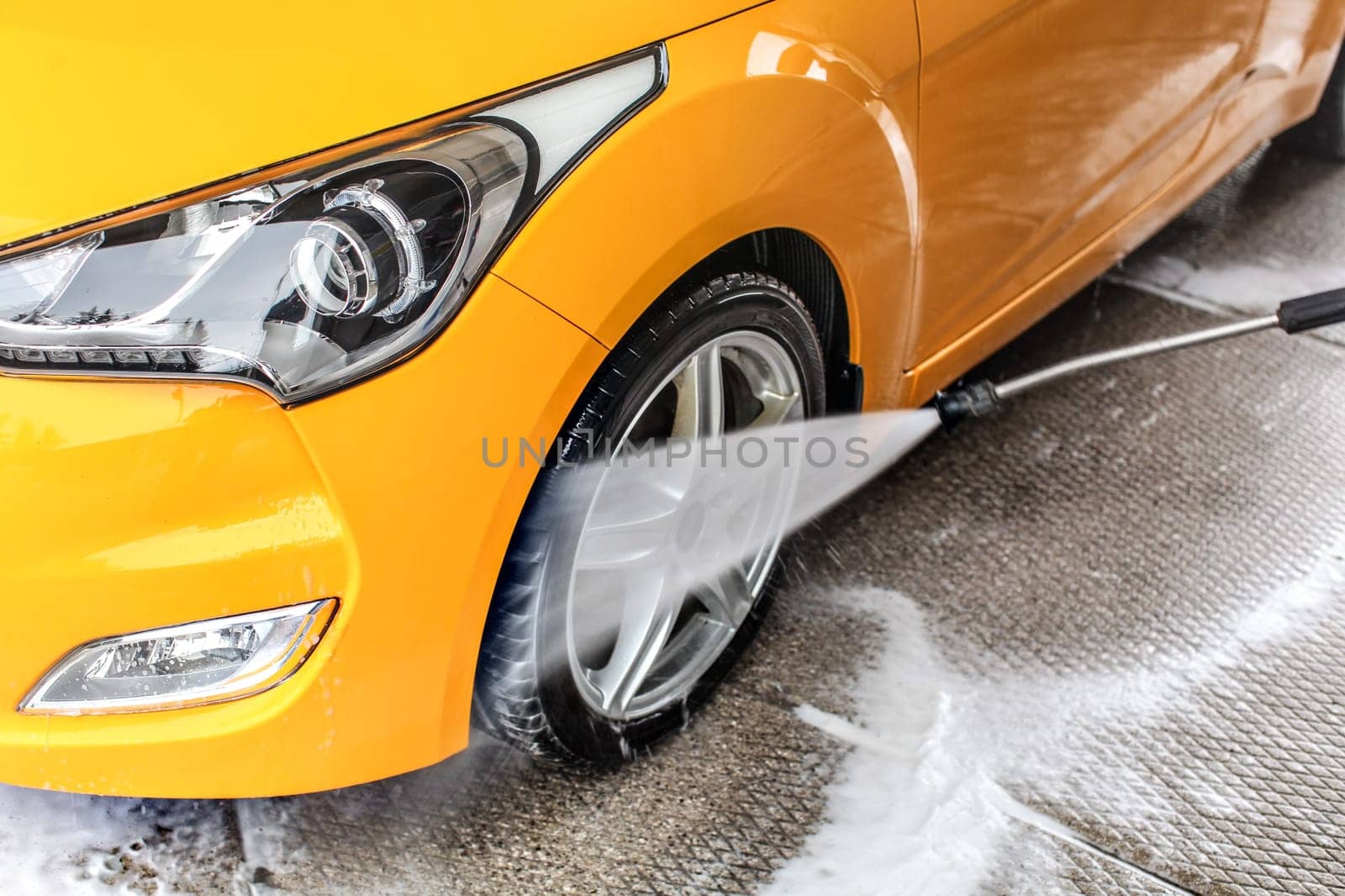 Detail on water jet spraying from the hose to yellow car front wheel and tyre, during cleaning in carwash.