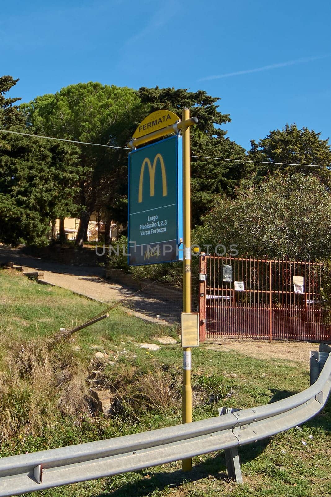 Civitavecchia, Italy - December 27, 2024: Bus stop sign with McDonald's logo and locations, in rural area with vegetation. Metal railing and red door in foreground. Concept: Branding in public spaces.