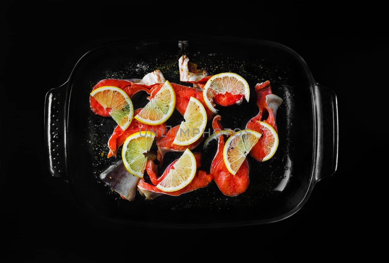 Pieces of red fish with lemon and seasoning in a glass dish on black background.