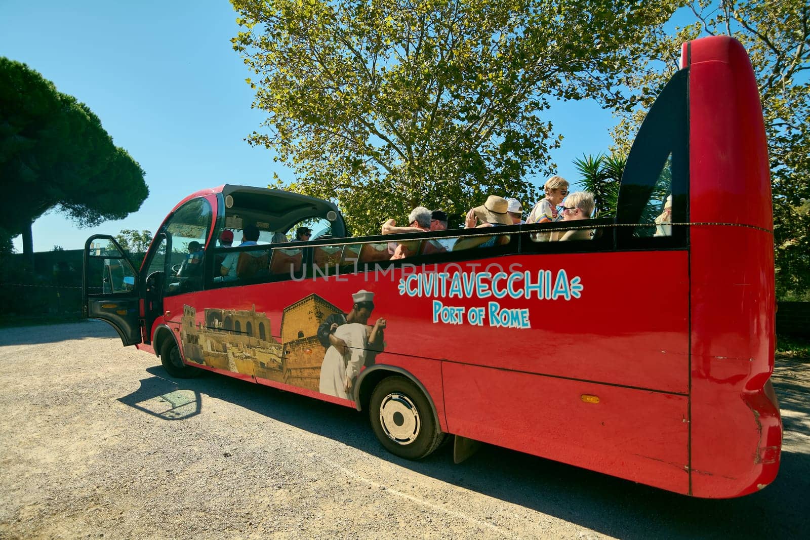 Civitavecchia. Italy - January 25, 2025: A vibrant red tourist bus with passengers, parked under a clear blue sky in Civitavecchia, Italy. The bus features images of local landmarks and the text Civitavecchia Port of Rome