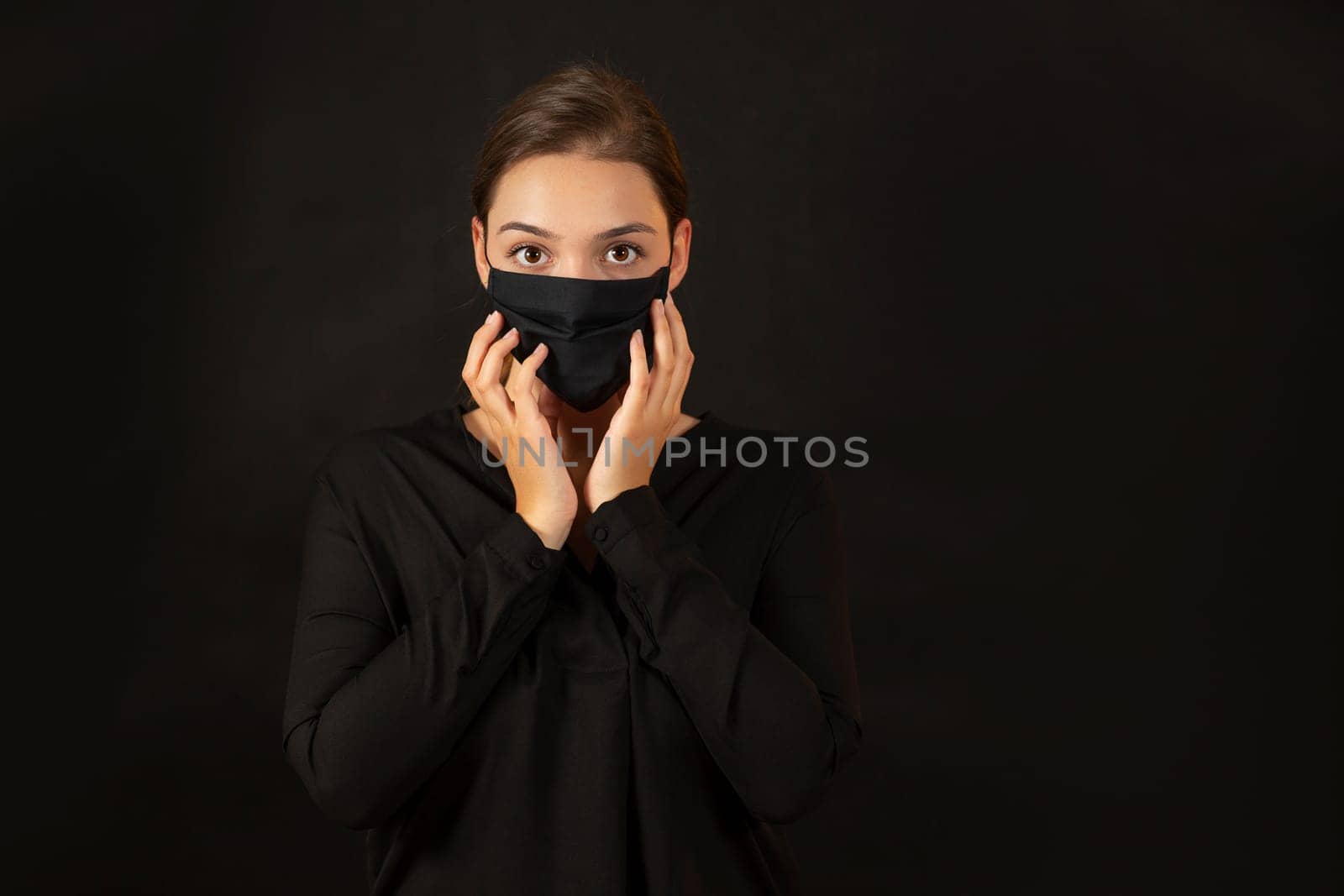 Attractive girl in black antiviral mask concerned about coronavirus prevention looking at the camera with copy space for text. Brunette young woman in studio touching her face.