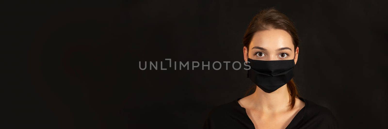 Horizontal head and shoulders portrait of a woman in protective mask amidst a Covid-19 pandemic. Studio headshot of attractive female against black backdrop with generous copyspace.
