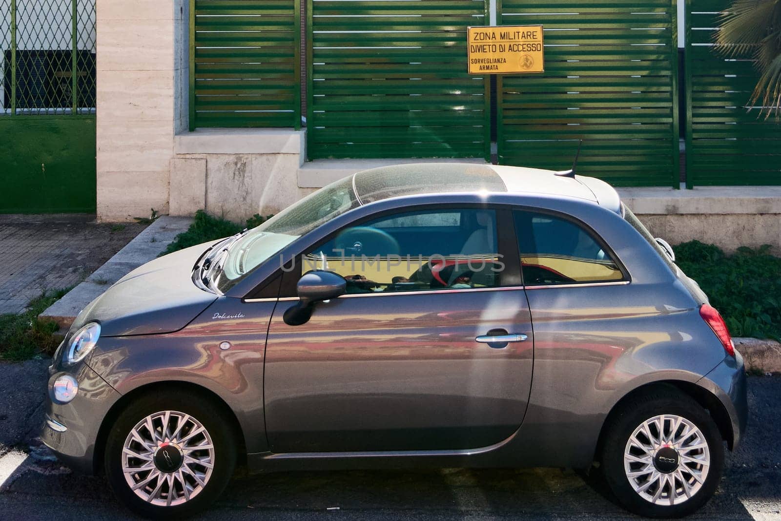 Civitavecchia, Italy - January 26, 2025: A small car parked next to a green fence with a yellow military zone sign, highlighting the contrast between the civilian vehicle and the restricted area.