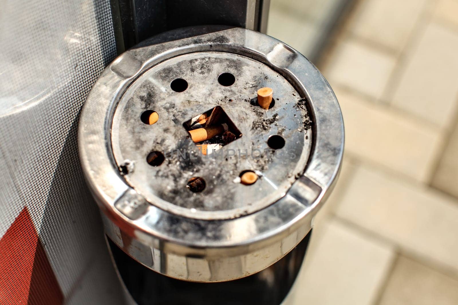 Sun shining on metal ashtray stand outside restaurant / office, with cigarette butts and ash. Smoking ban inside concept.