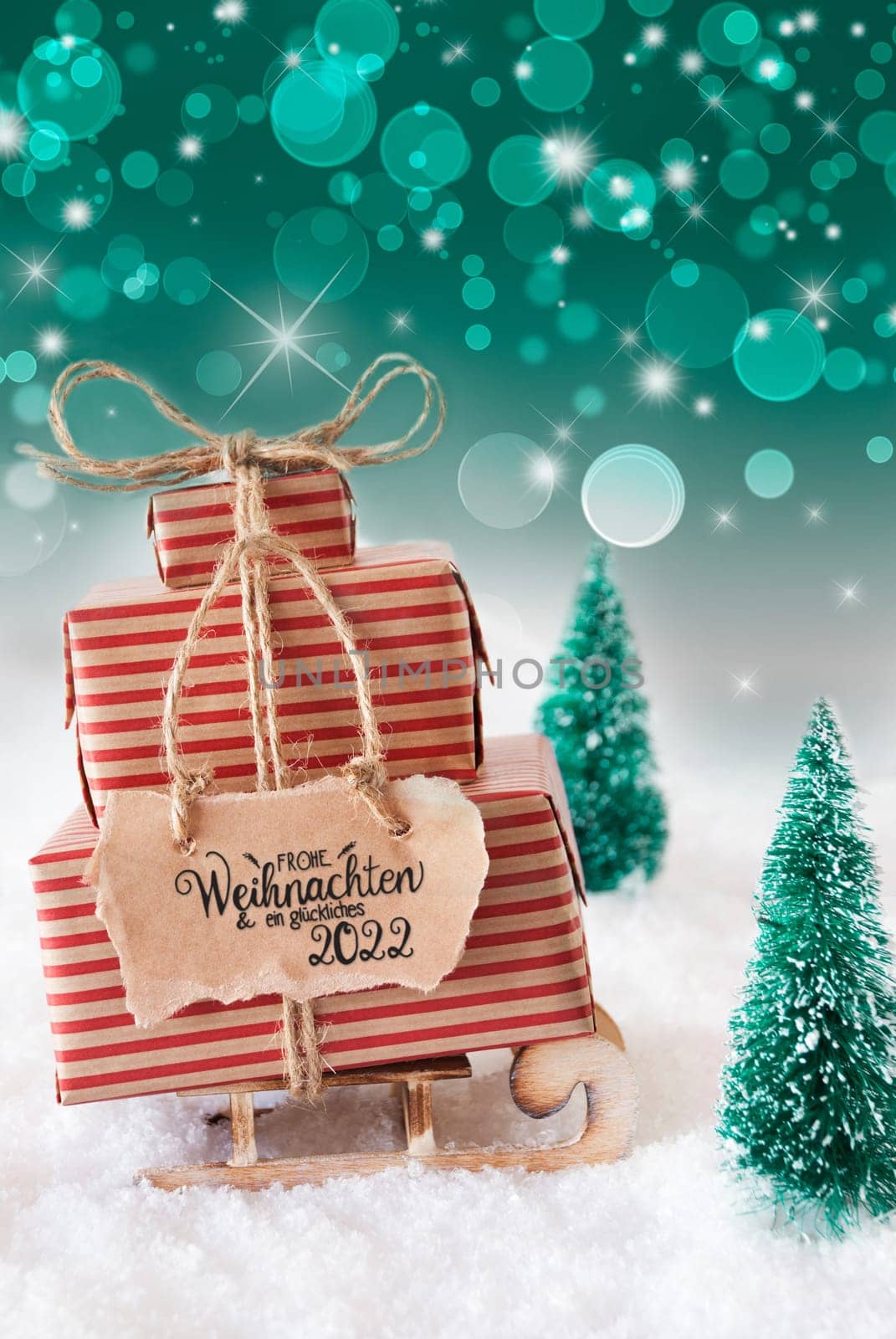 Sled With Christmas Gift And Label With German Frohe Weihnachten Und Ein Glueckliches 2022 Means Merry Christmas And A Happy 2022. Snow With Christmas Tree And Green Sparkling Background