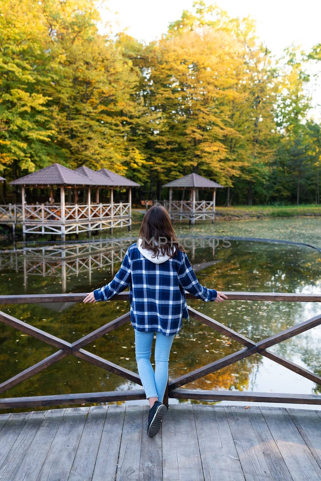 A young brunette woman in a plaid shirt stands with her back near the pond and enjoys the autumn warm day