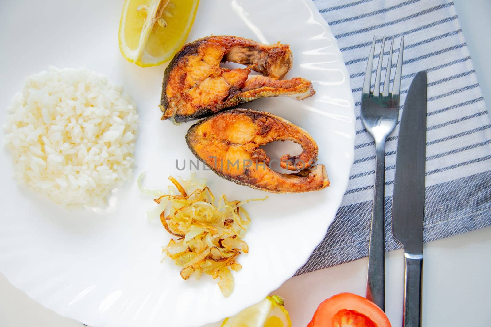 Fried fish with onions, rice and lemon lies on a plate, on a waffle towel and cutlery on a white background. High quality photo