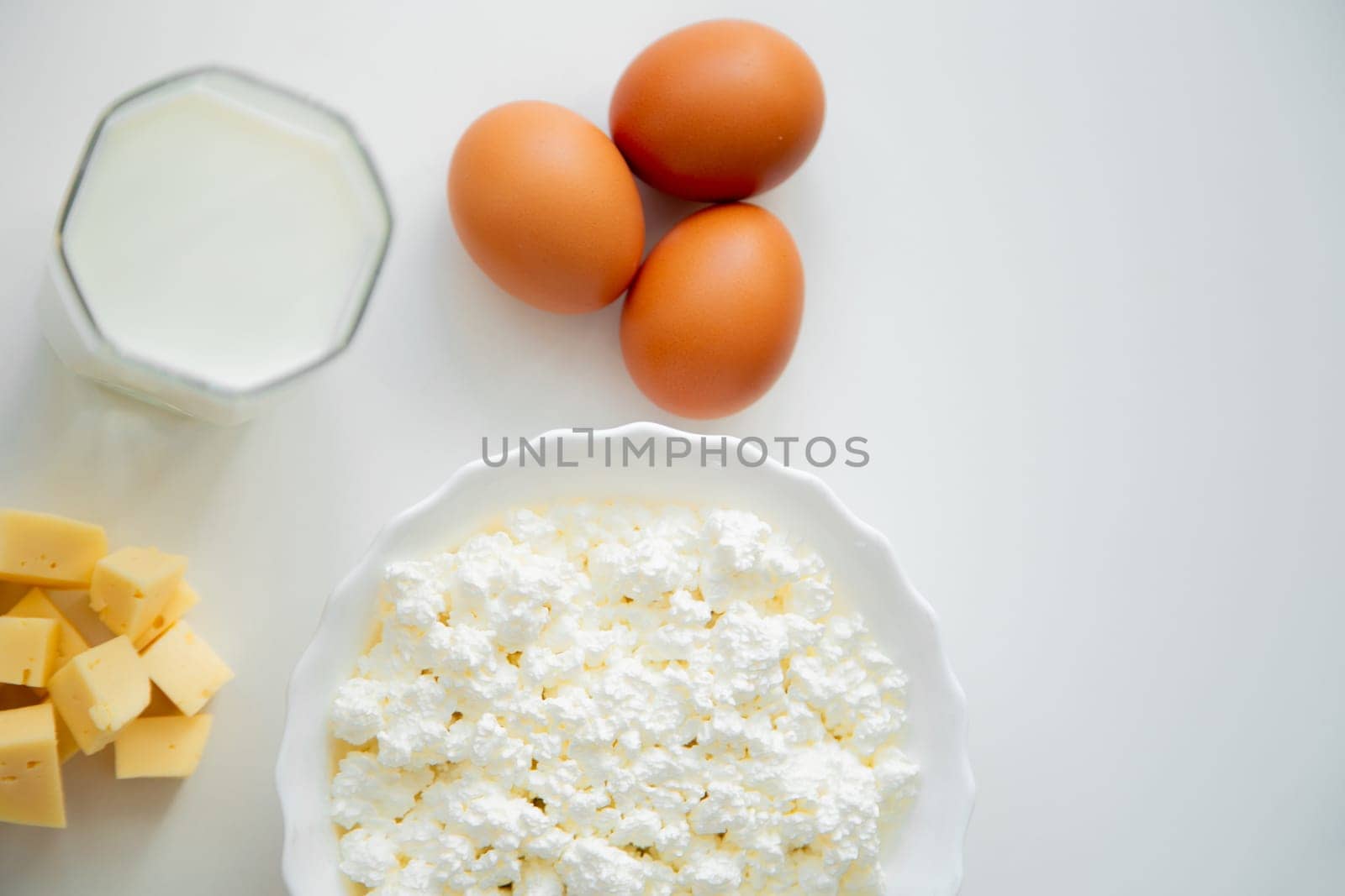 A white bowl with cottage cheese, eggs, diced cheese and a glass of milk stand on a white background. . High quality photo