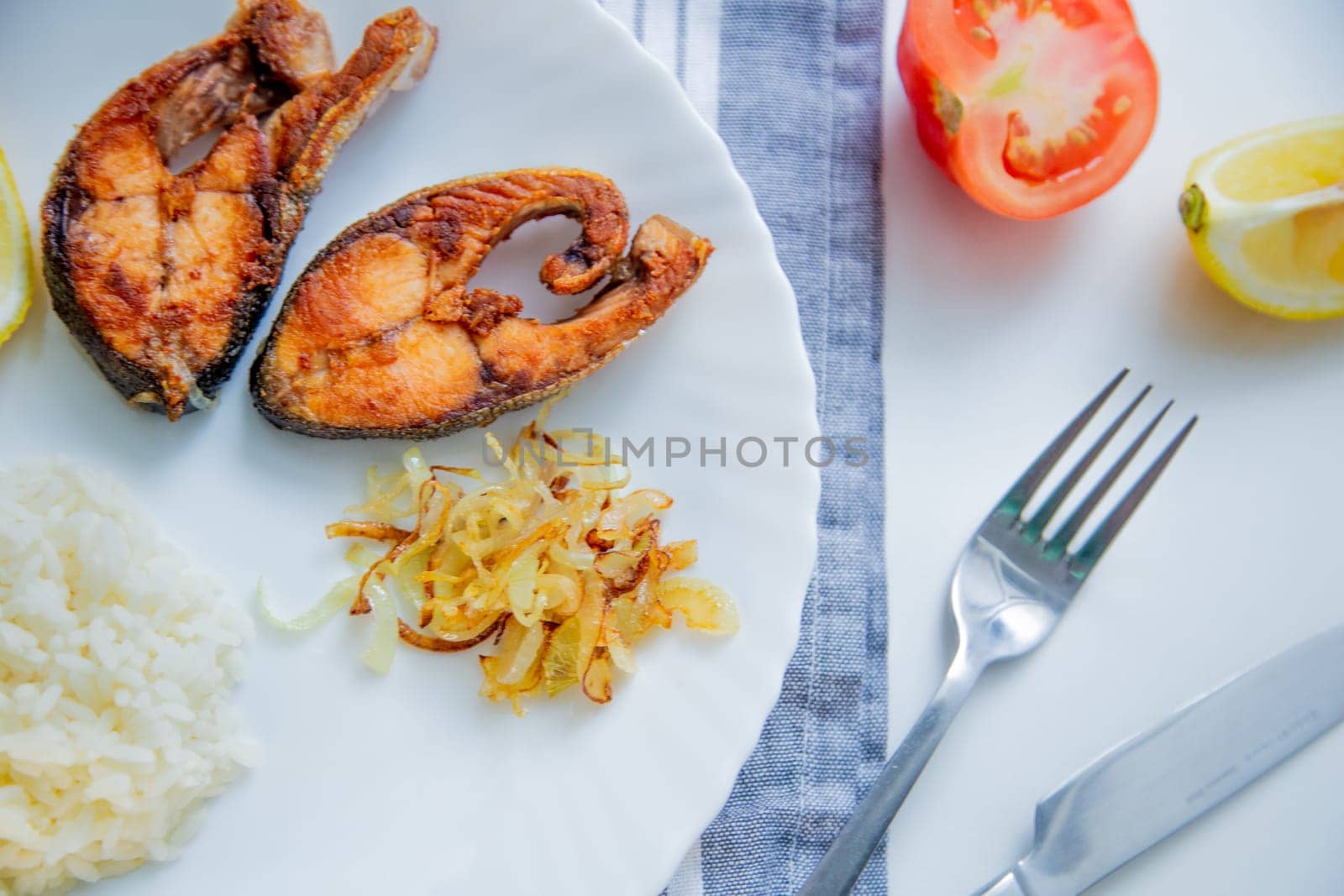 Fried fish with onions, rice and lemon lies on a plate, on a waffle towel and cutlery on a white background. High quality photo