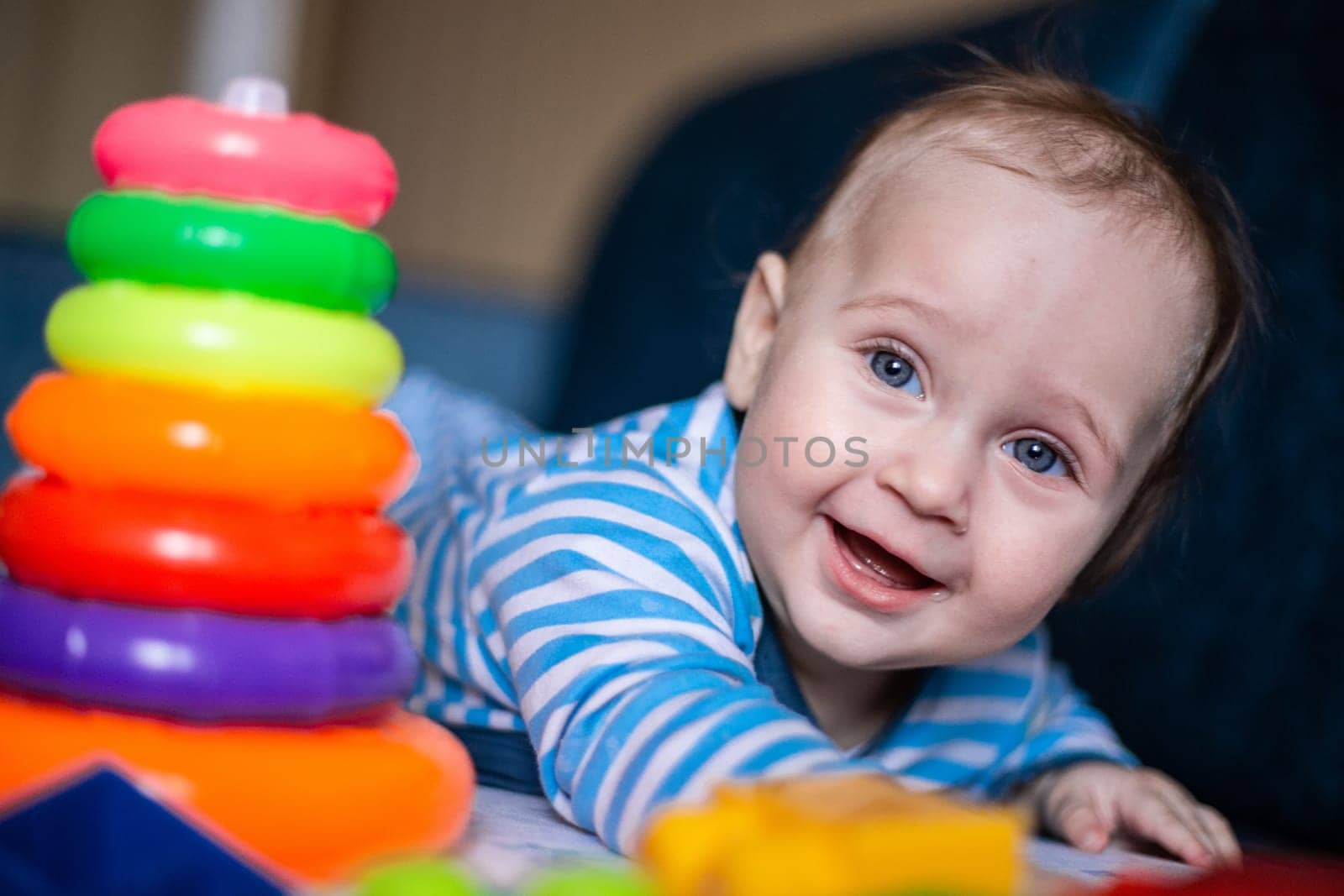 A little kid in a striped suit is lying on his tummy, next to him a children's pyramid and toys. High quality photo