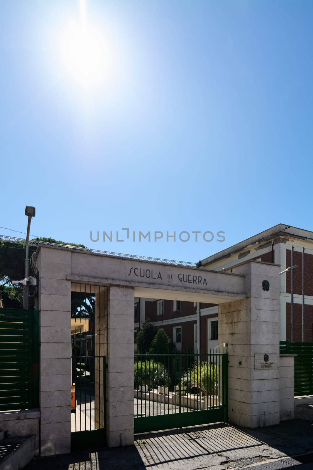 Civitavecchia. Italy - January 26, 2025: The entrance gate of the Italian War School in Civitavecchia, with a sign, green metal fence, and a red brick building, under a bright sunny sky.