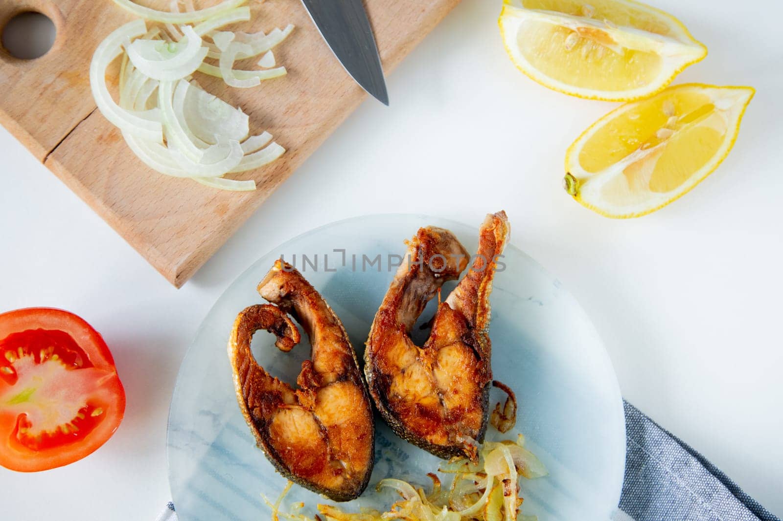 Fried fish with onions is on a plate, on a waffle towel, next to it is a cutting board with chopped onions, lemon and tomato on a white background. High quality photo