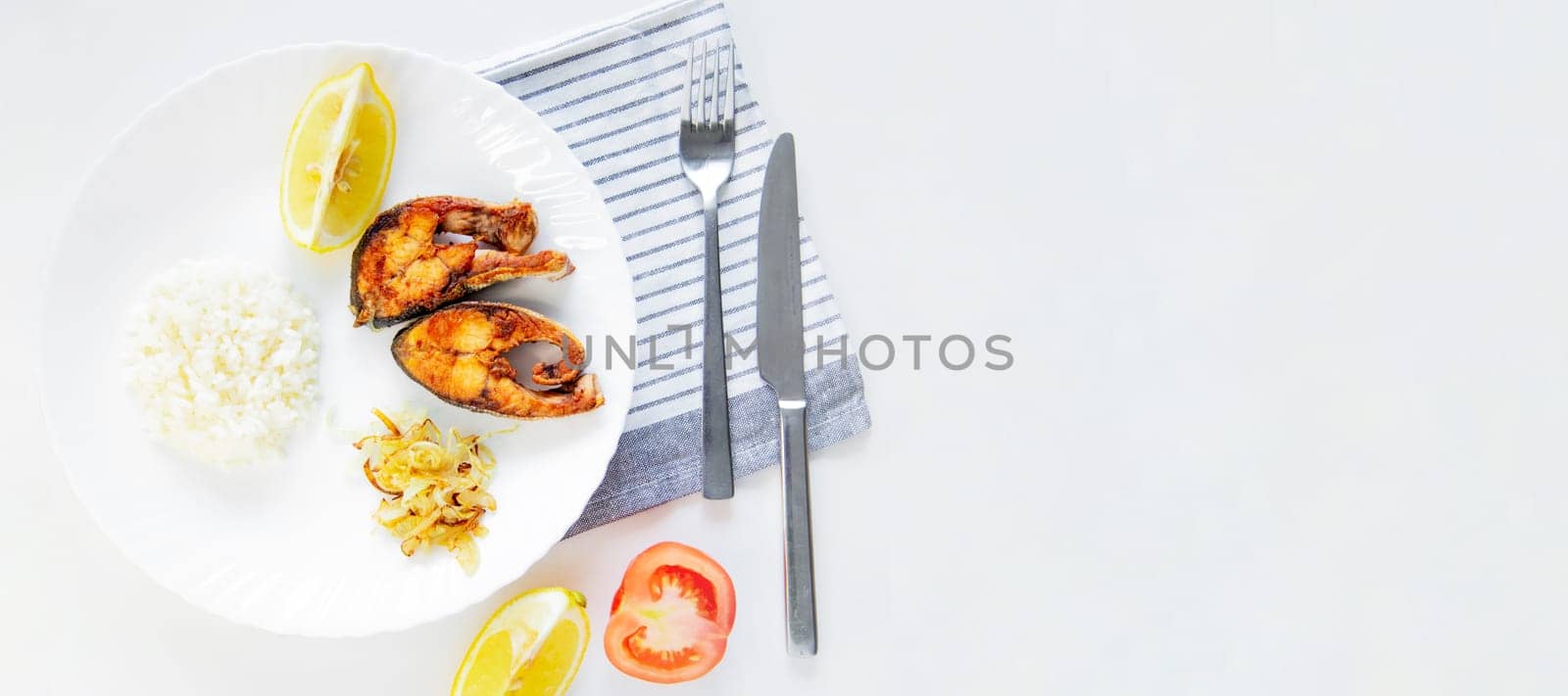 Fried fish with onions, rice and lemon lies on a plate, on a waffle towel and cutlery on a white background. High quality photo