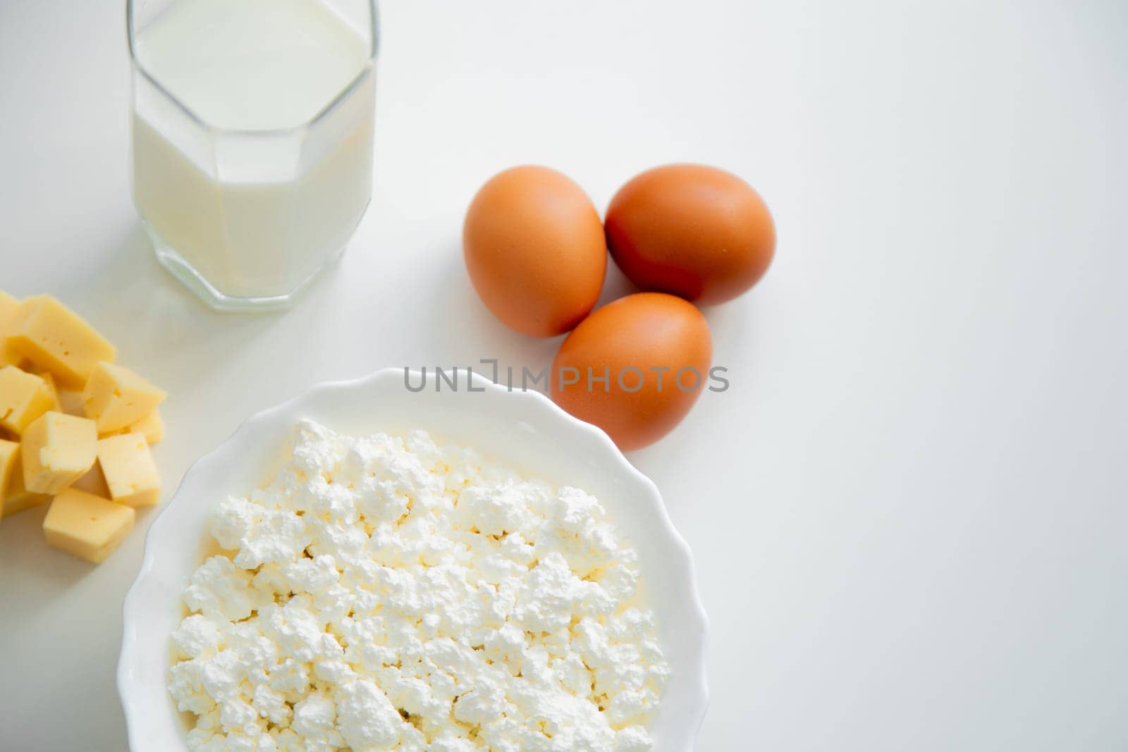 A white bowl with cottage cheese, eggs, diced cheese and a glass of milk stand on a white background. . High quality photo