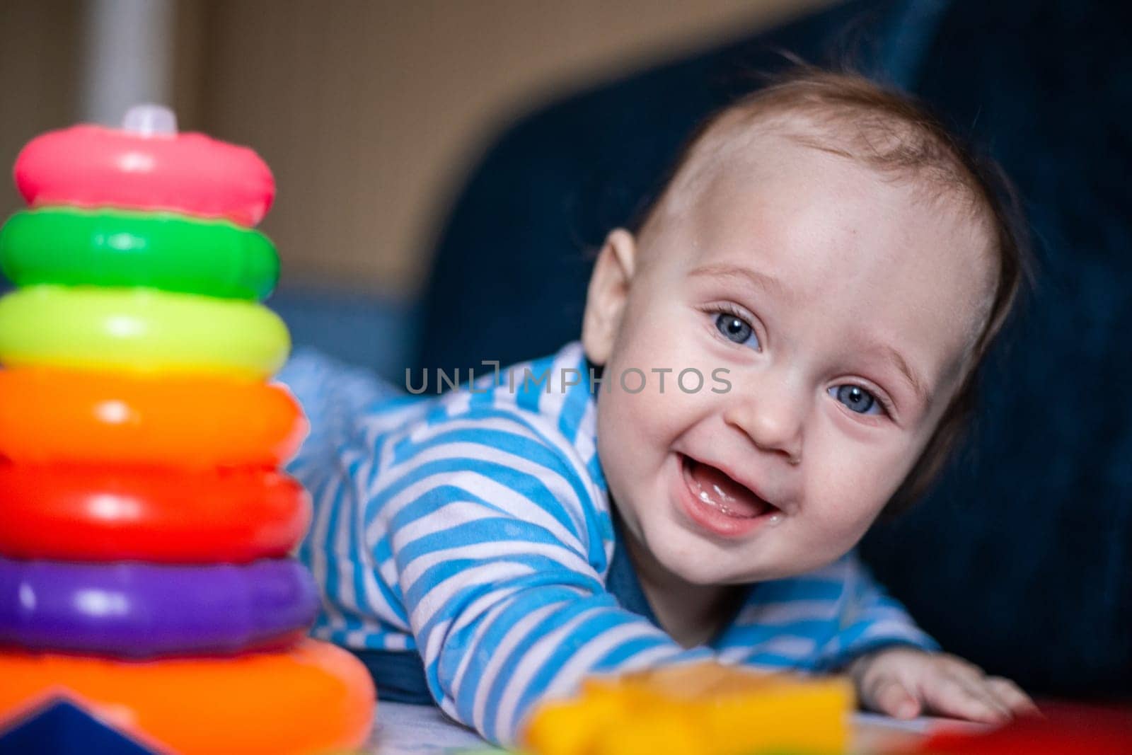 A little kid in a striped suit is lying on his tummy, next to him a children's pyramid and toys. High quality photo