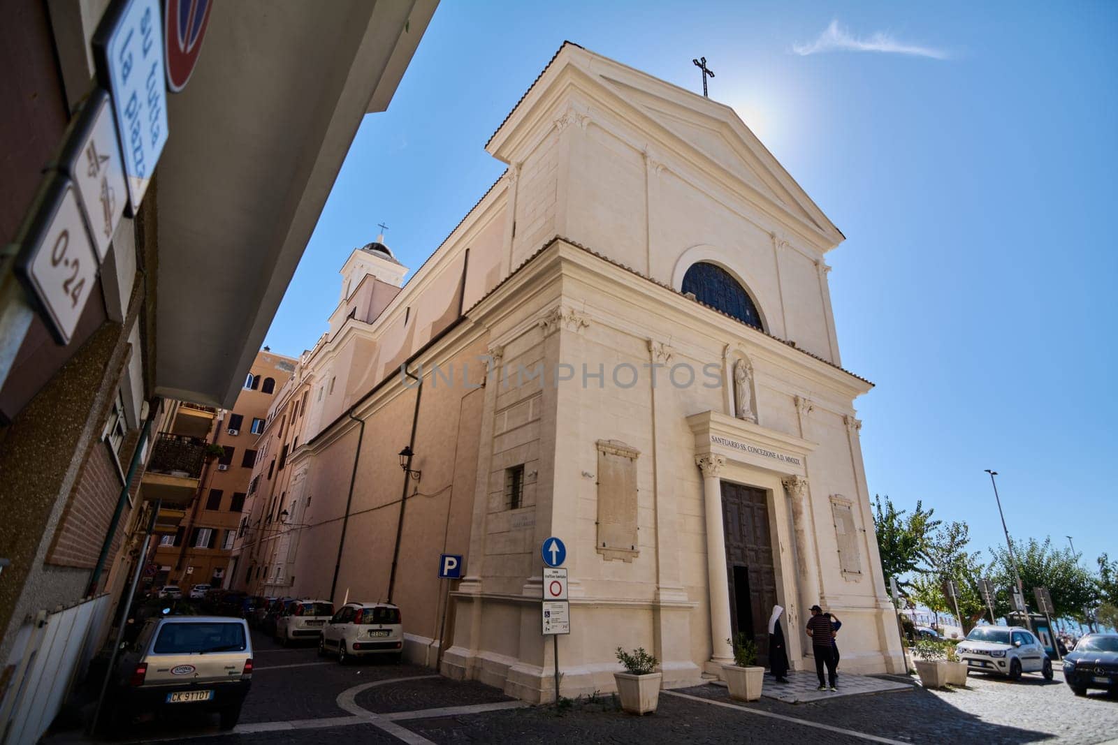 Civitavecchia, Italy - January 27, 2025: Sunlit church in Civitavecchia, Italy. The historic building, people entering, parked cars and blue sky create a vibrant scene of Italian life and architecture
