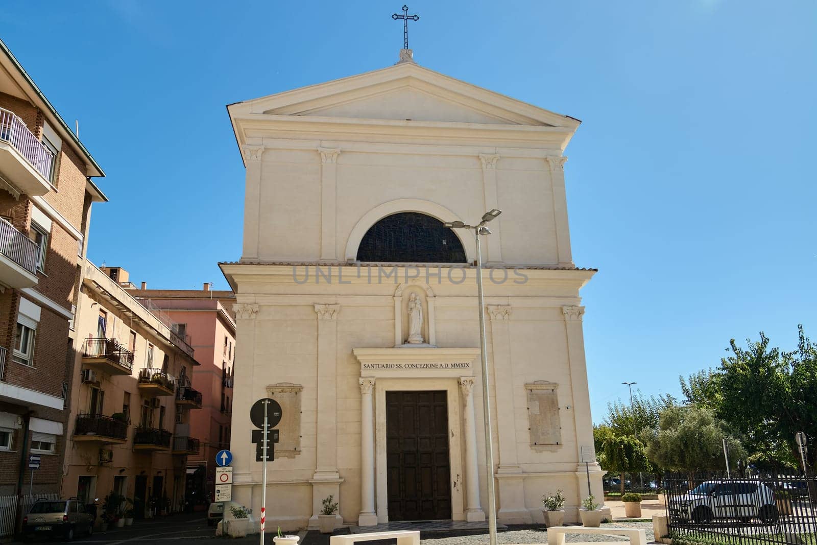 Civitavecchia church entrance with people entering. The sunlit facade, parked cars, and blue sky create a picturesque scene of Italian architecture and daily life.