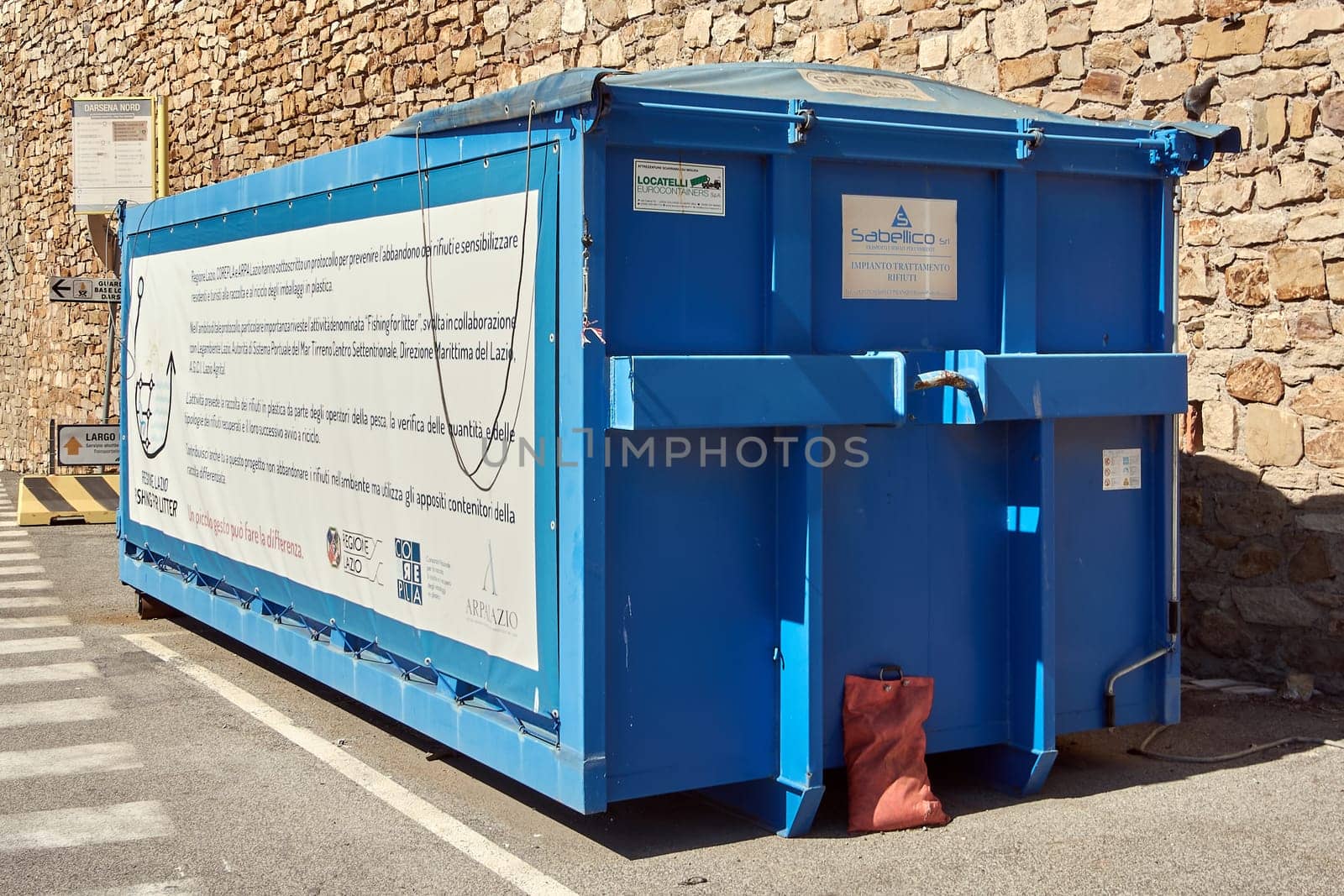 Civitavecchia. Italy - January 31, 2025: Large blue waste container in an urban setting against a stone wall. The container has various labels and signs, indicating its use for waste management.