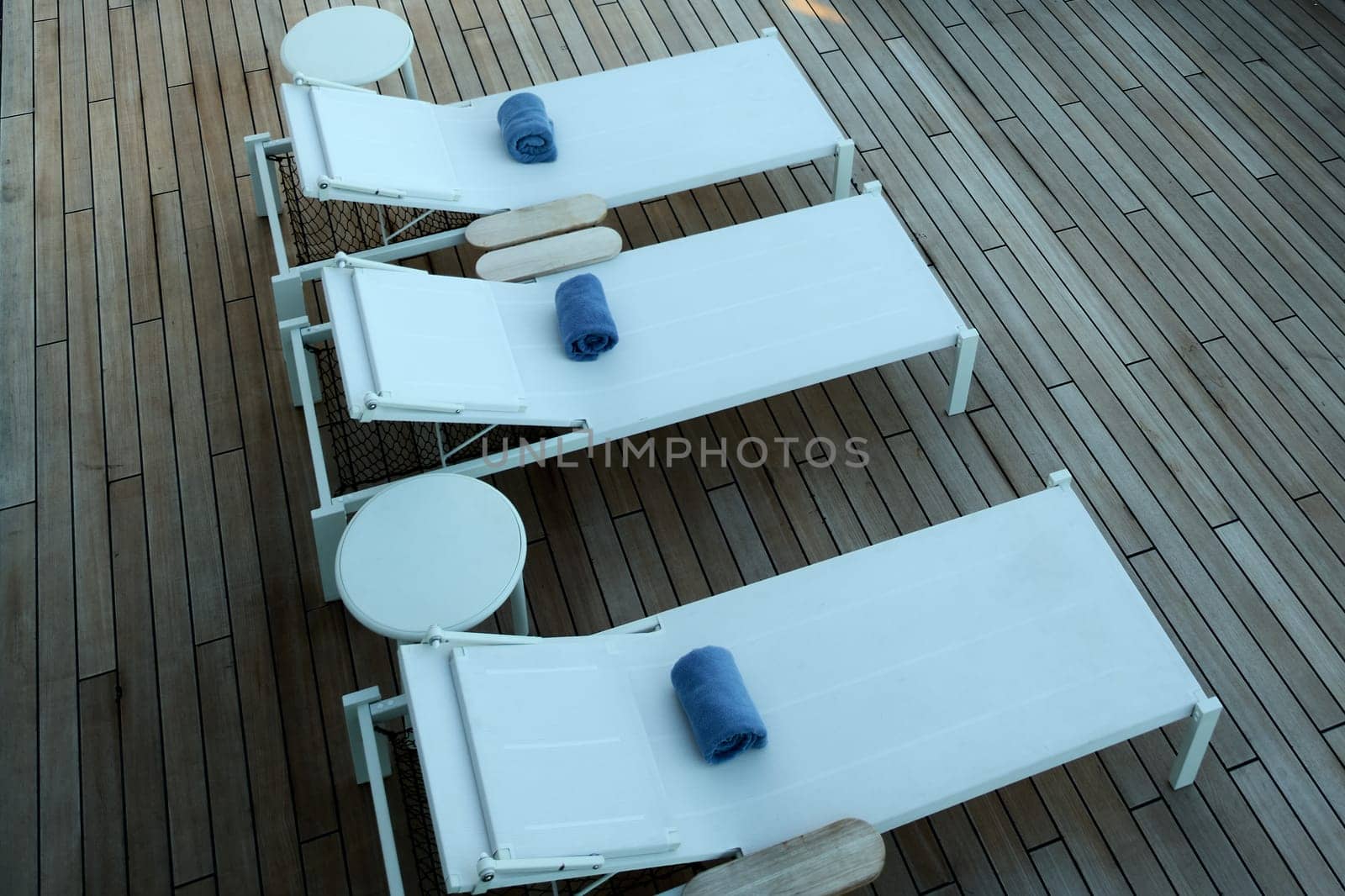 Relaxation. Three white sun loungers with neatly rolled blue towels on a wooden deck. A peaceful outdoor setting ideal for travel, leisure, or wellness concepts.