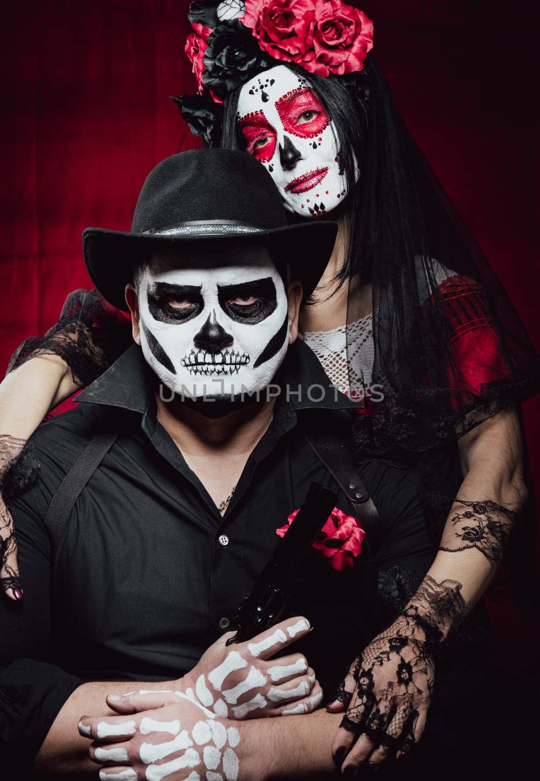 beautiful woman with a sugar skull makeup with a wreath of flowers on her head and a skeleton man in a black hat holding a gun