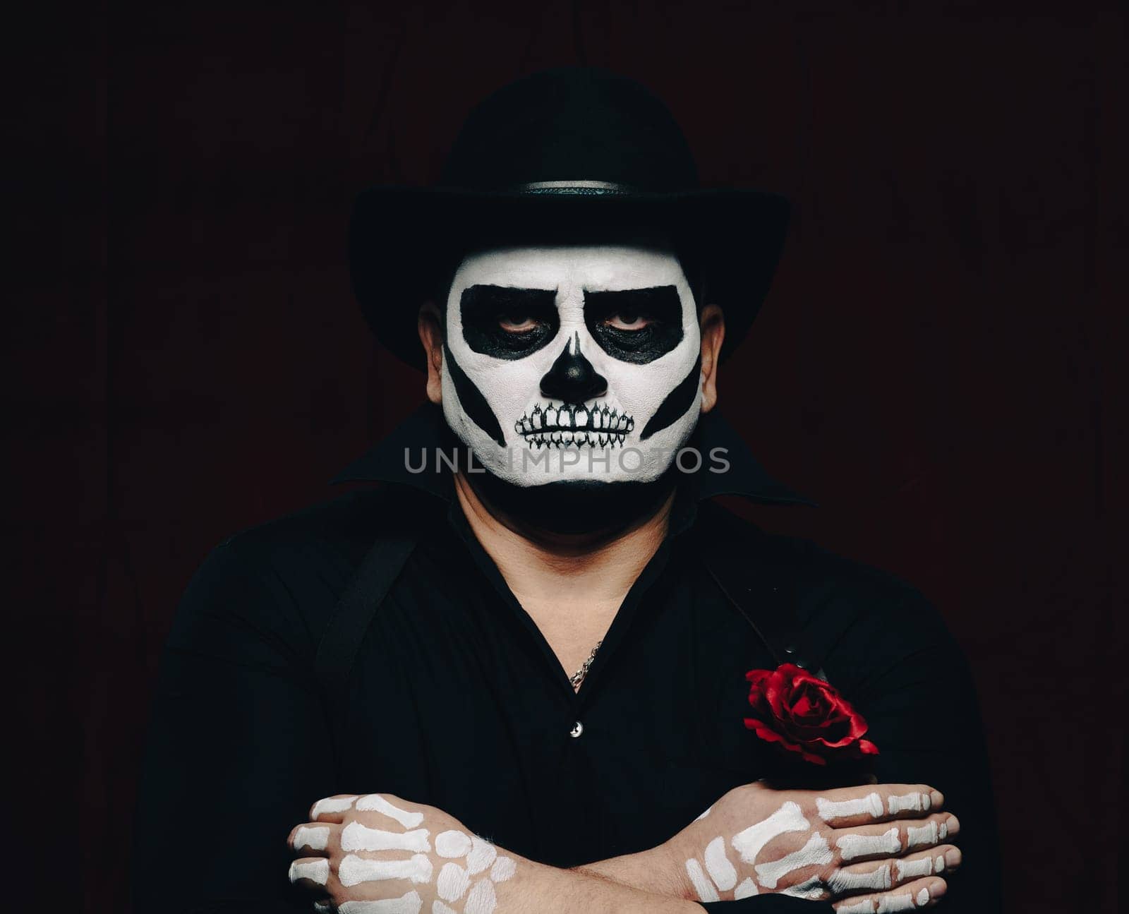 a man is painted with a skeleton in a black hat and shirt, arms crossed on his chest. Black background