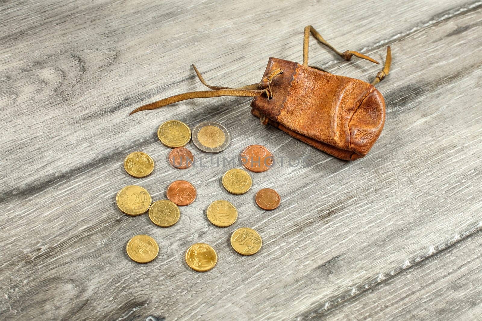Old worn brown leather coin pouch,  with euro coins spilled on gray wood desk.