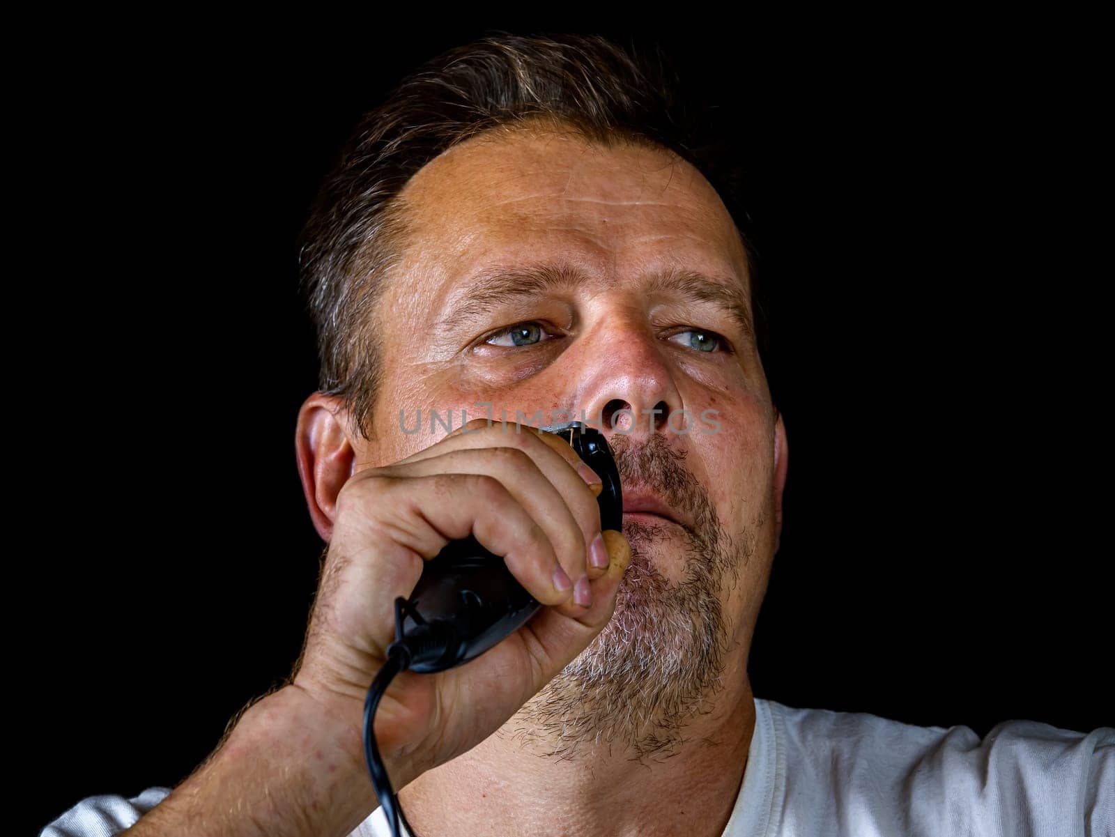 A middle-aged man cuts his gray beard with a shaving trimmer. Mustache and beard clipper. Adult. Male hairdresser. Man's face. Aged people. Hairdresser's tool. Facial care.