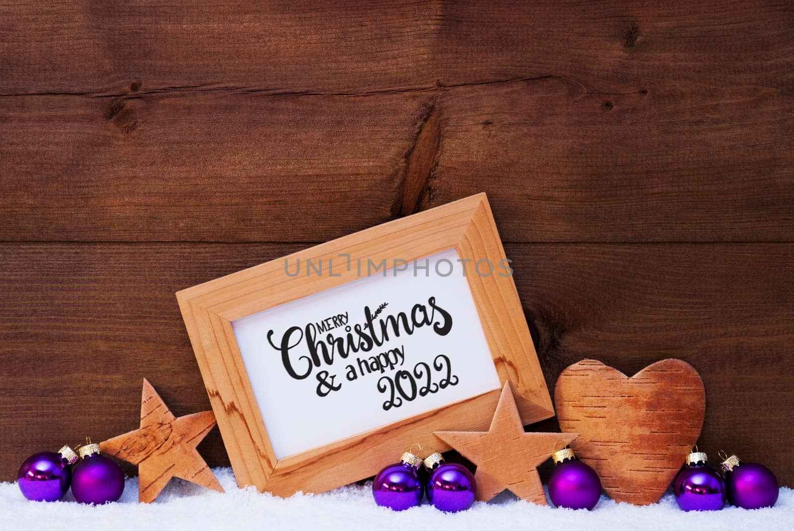 Frame With English Calligraphy Merry Christmas And A Happy 2022. Purple Christmas Decoration Like Ball, Star And Heart. Wooden Background With Snow