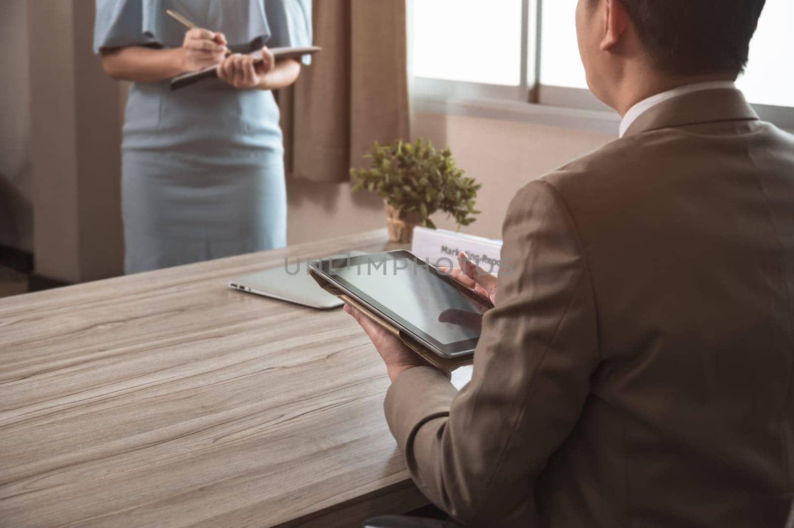 Businessman manager in brown suit using PC tablet while sitting at office desk and secretary in blue dress taking notes while standing in front of his boss. Business work concept