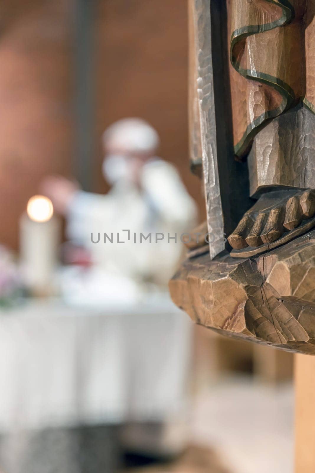 Carved wooden feet, detail of wooden statue of a Saint. In the blurred background a priest celebrates Mass.
