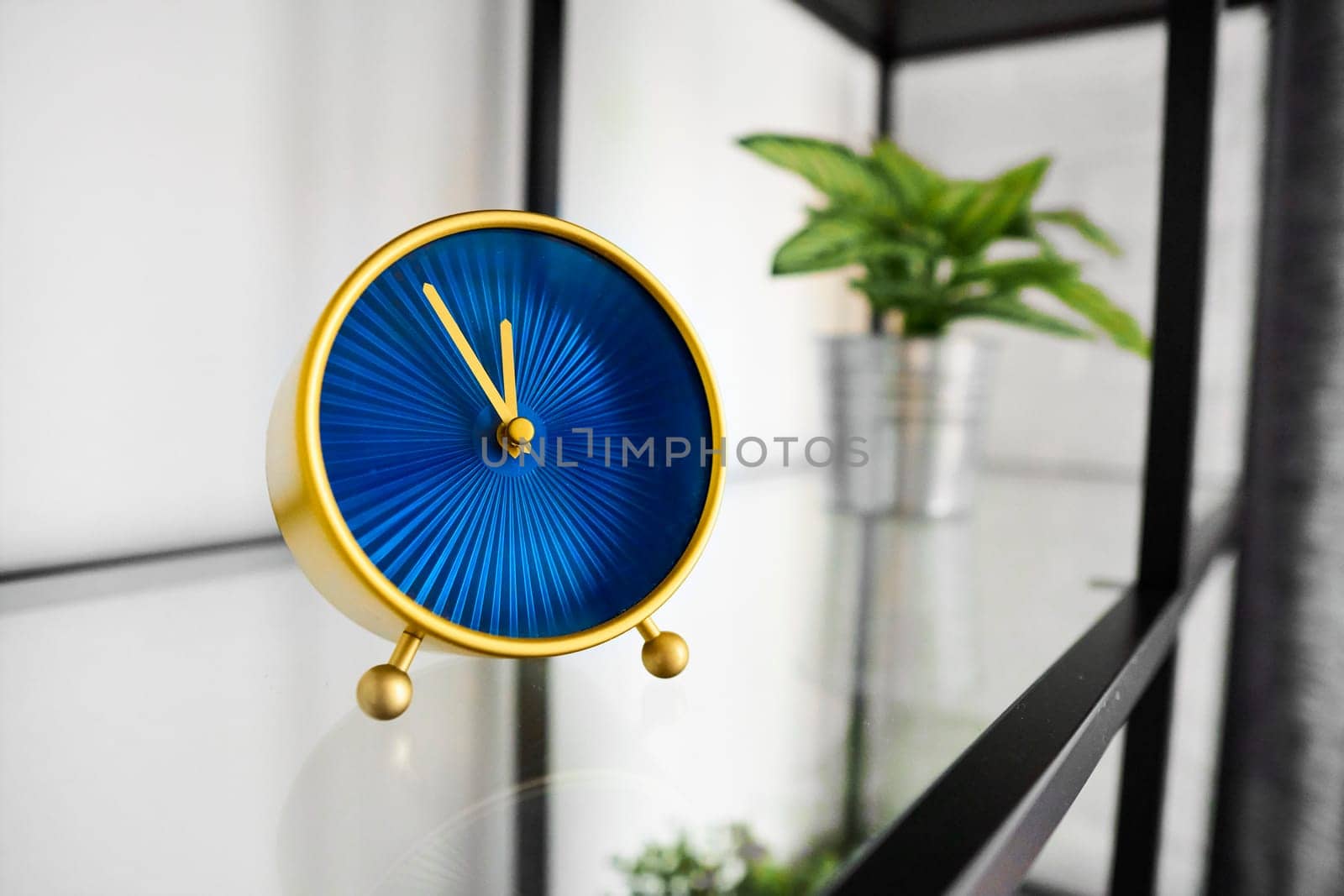 Small golden tabletop clock with blue analog face on glass shelf. Popular round clock with hour and minute hands on glossy surface in office. Decorative mantel clock on rack at home. Quartz movement