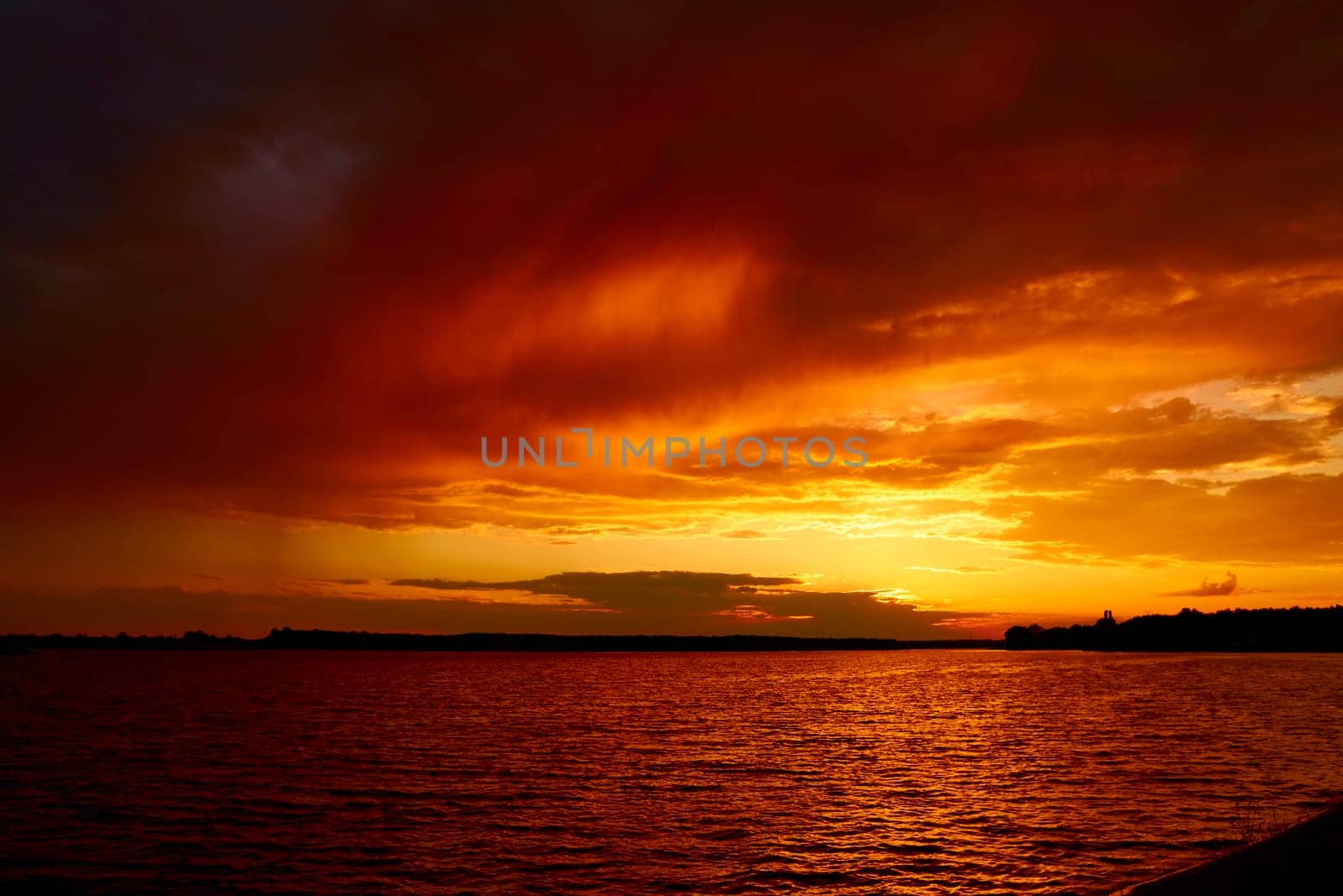 Bloody red cloudy sunset above lake. Morning fiery orange sunrise over river. Reflective water surface waves under clouds at dusk in summer. Evening cloudscape in twilight. Ruddy horizon sky and river