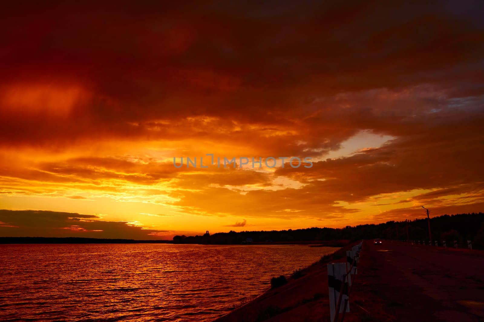 Red cloudy sunset above lake. Last day on Earth. The end of world. Apocalypse concept. Fiery orange sunrise over river dam. Car on the road under clouds at dusk. Cloudscape in twilight. Bloody sky