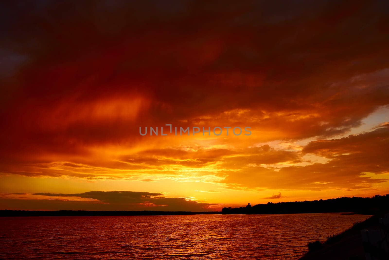 Red cloudy sunset above lake at dusk. Bloody sky above reflective water surface waves in wind. Evening cloudscape in twilight. Ruddy horizon sky and river. Morning fiery orange sunrise in summer dawn