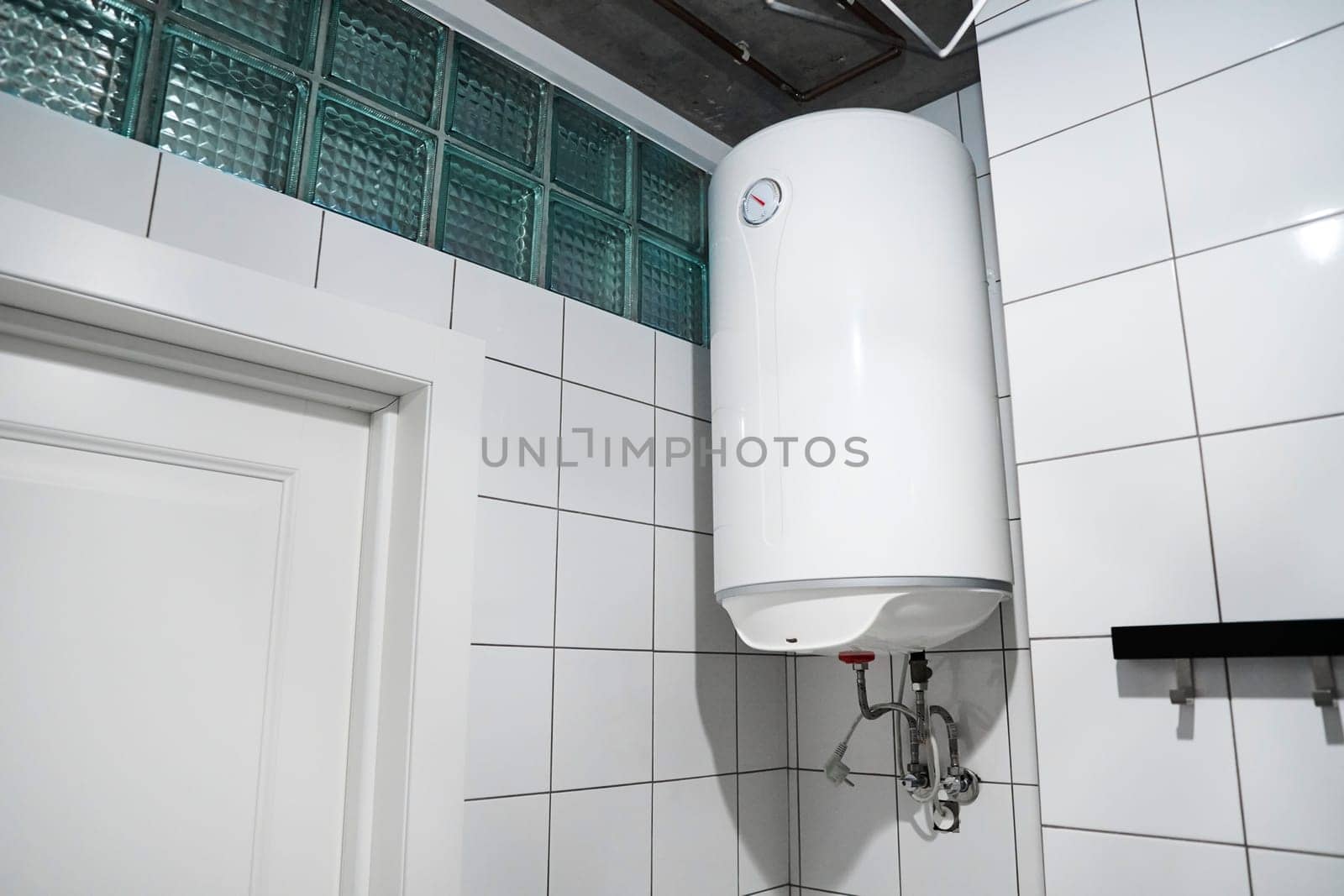 Household budget water heater hanging on the wall in boiler room. Modern gas tanked boiler in bathroom. Сommon electric storage tank water heater. Home heating system in front of white tiled wall