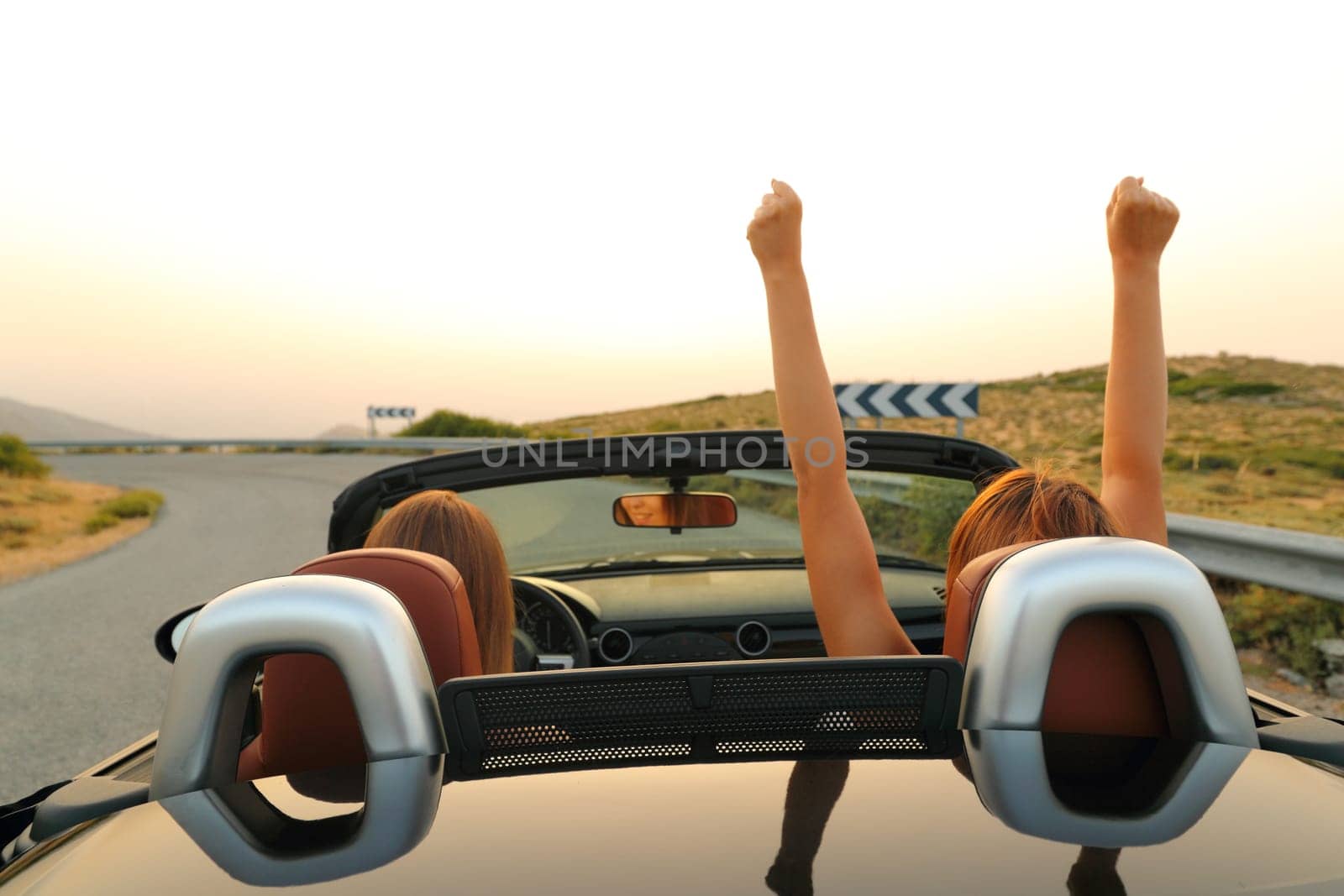 Back view of two excited women driving a convertible car in the road on summer vacation