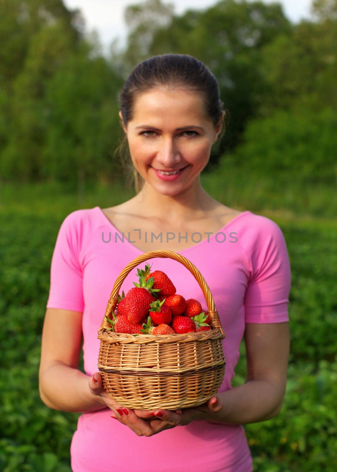 Young woman in pink holding basket full of strawberries just picked from the field behind her. Shallow depth of field - focus on strawberry only